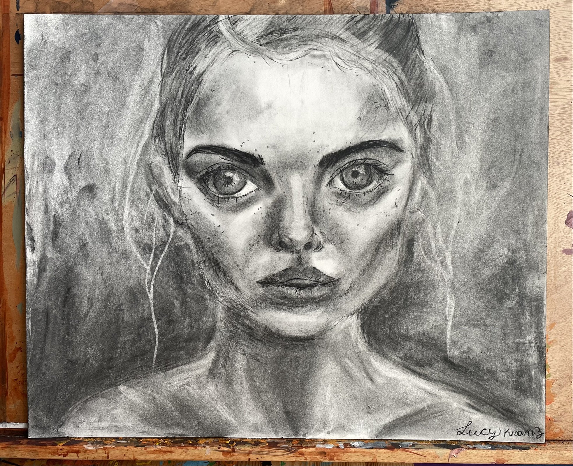 A grayscale portrait drawing of a woman with large eyes, slightly parted lips, and long hair. The drawing emphasizes her facial features with detailed shading and has a realistic yet stylized appearance. The artist's signature, Lucy Krantz, is in the