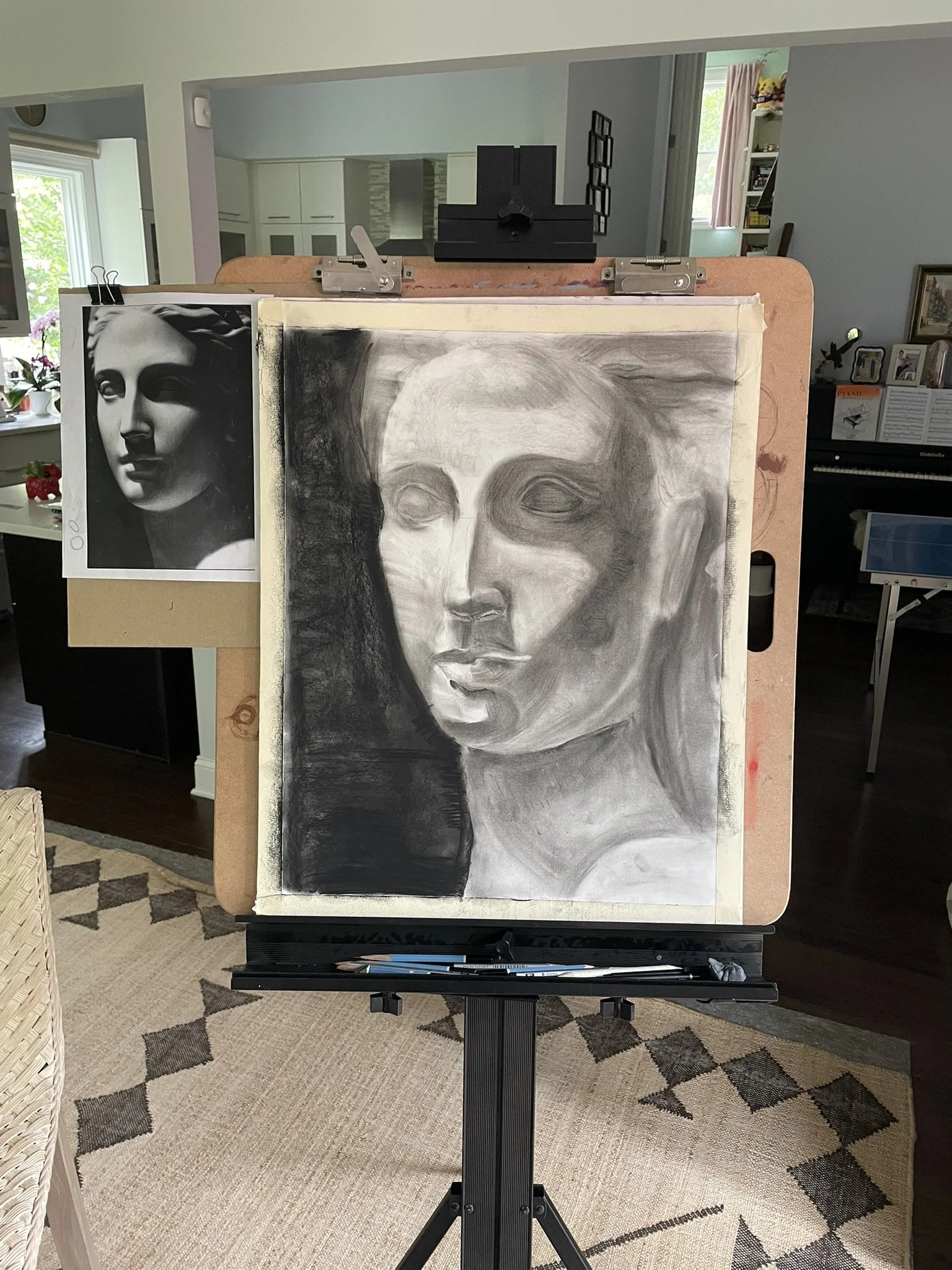 An art studio with an unfinished charcoal portrait of a woman on an easel and a reference photo of a sculpture of a woman's face attached to the left side of the easel.