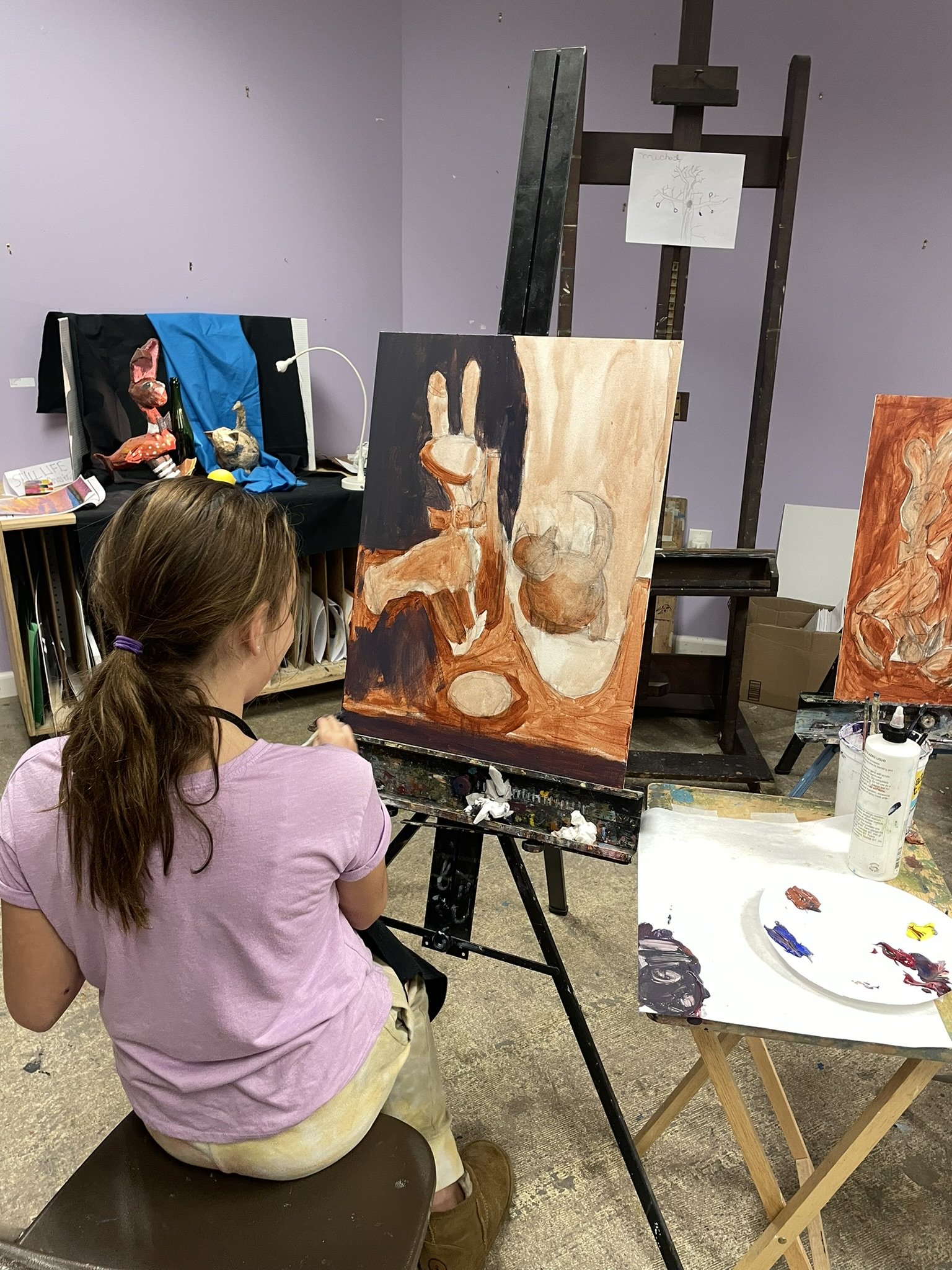 A young girl with brown hair tied back, wearing a pink t-shirt, sitting on a brown bench and painting a still life on a canvas in an art studio. The painting depicts a ceramic orange teapot and other objects on a table. The studio has a purple wall, 