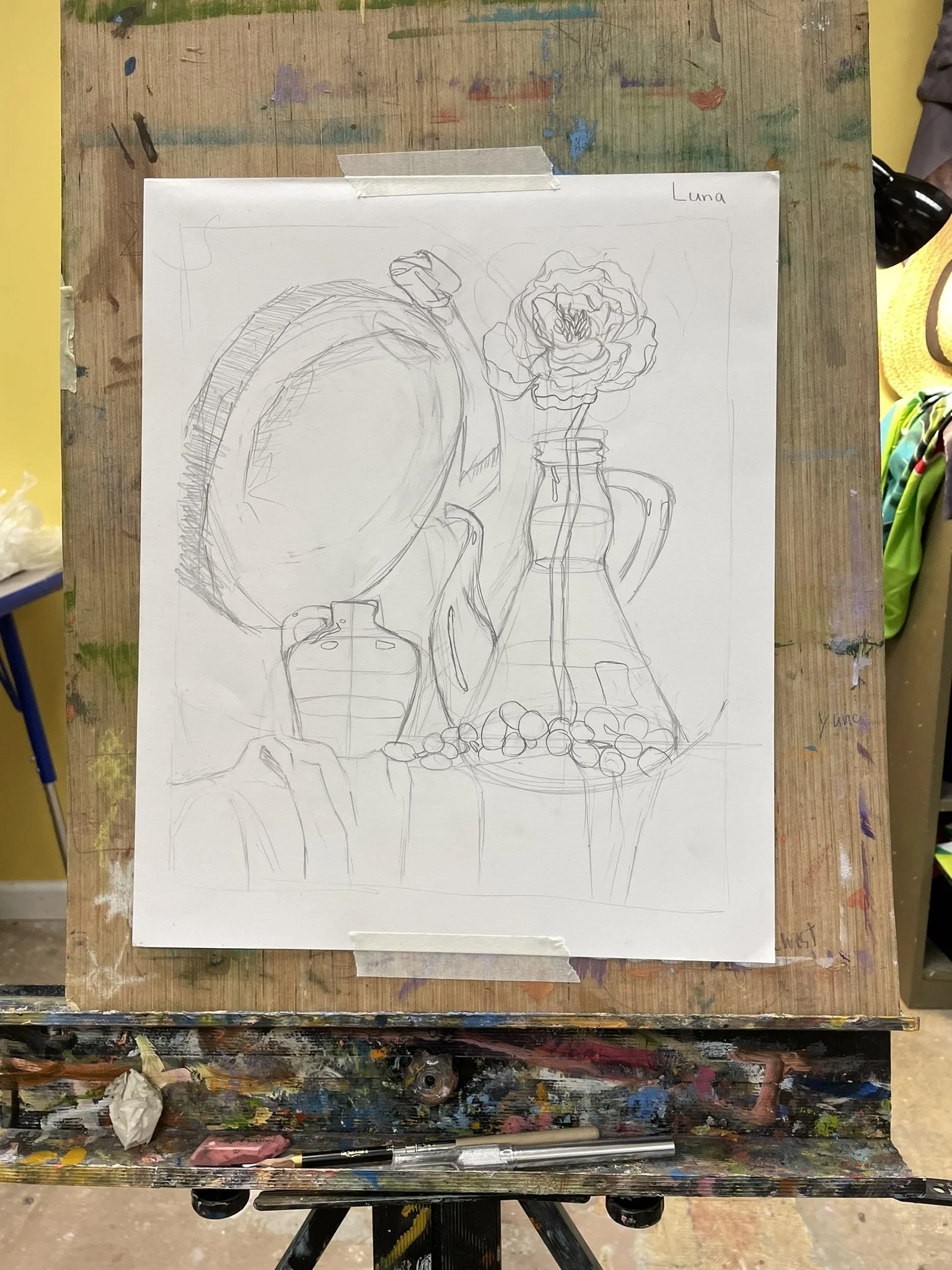 A pencil sketch on a white paper taped to a colorful, paint-stained wooden easel. The sketch depicts a still life arrangement with a large round object, a flower in a vase, bottles, and scattered small objects. The top right corner of the paper has t