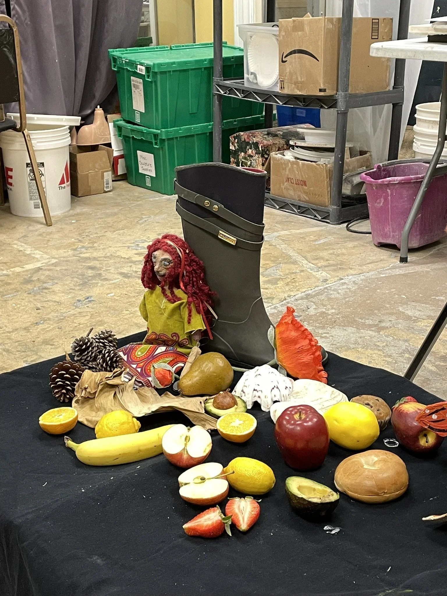 A table with various fruits and a doll with red yarn hair, positioned in front of a black boot-shaped object, in a storage or workshop area.