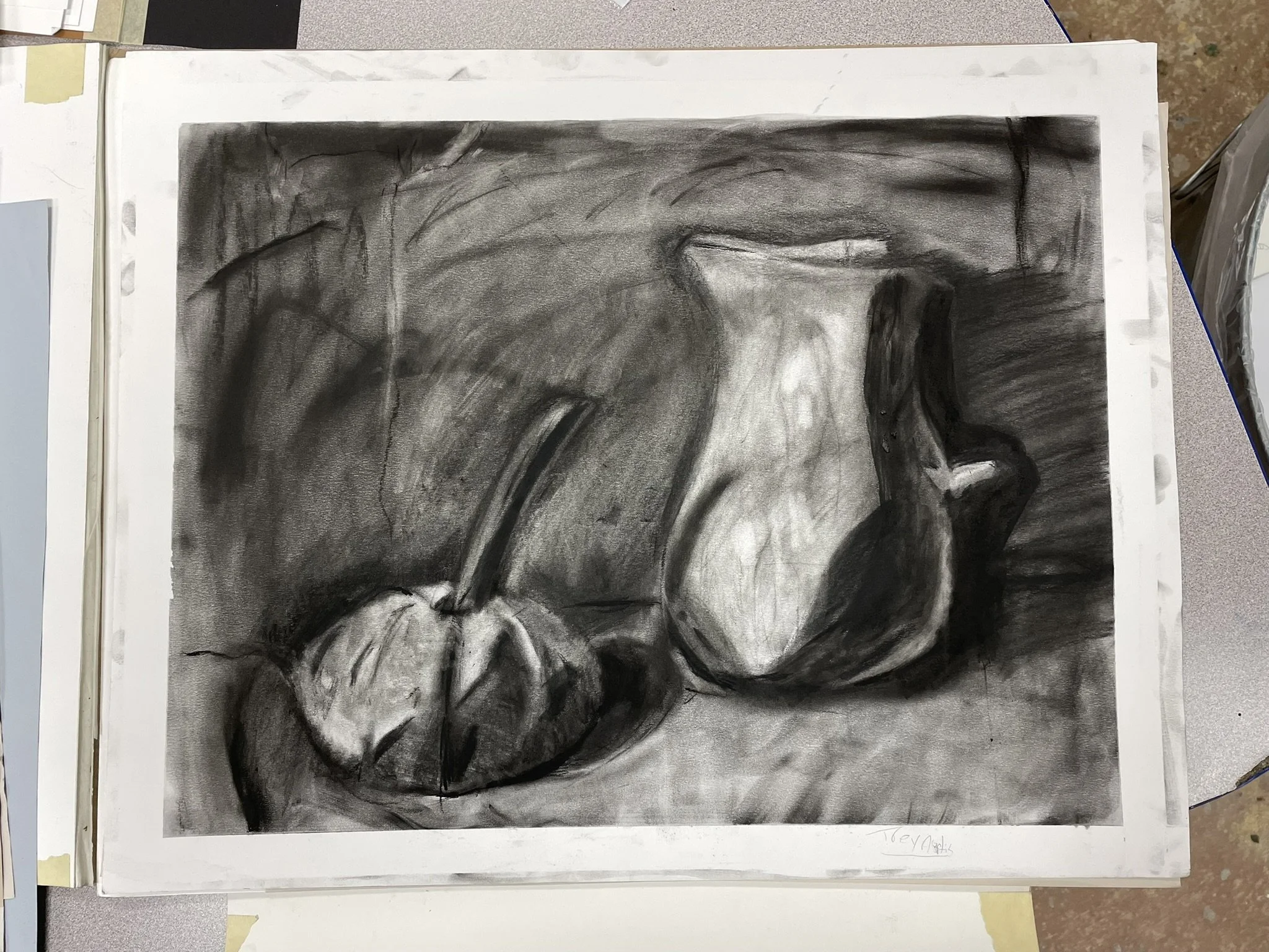 A black and white charcoal drawing of two pears placed on a surface. One pear is whole and the other is sliced open, revealing the inside. The background is shaded, highlighting the pears.