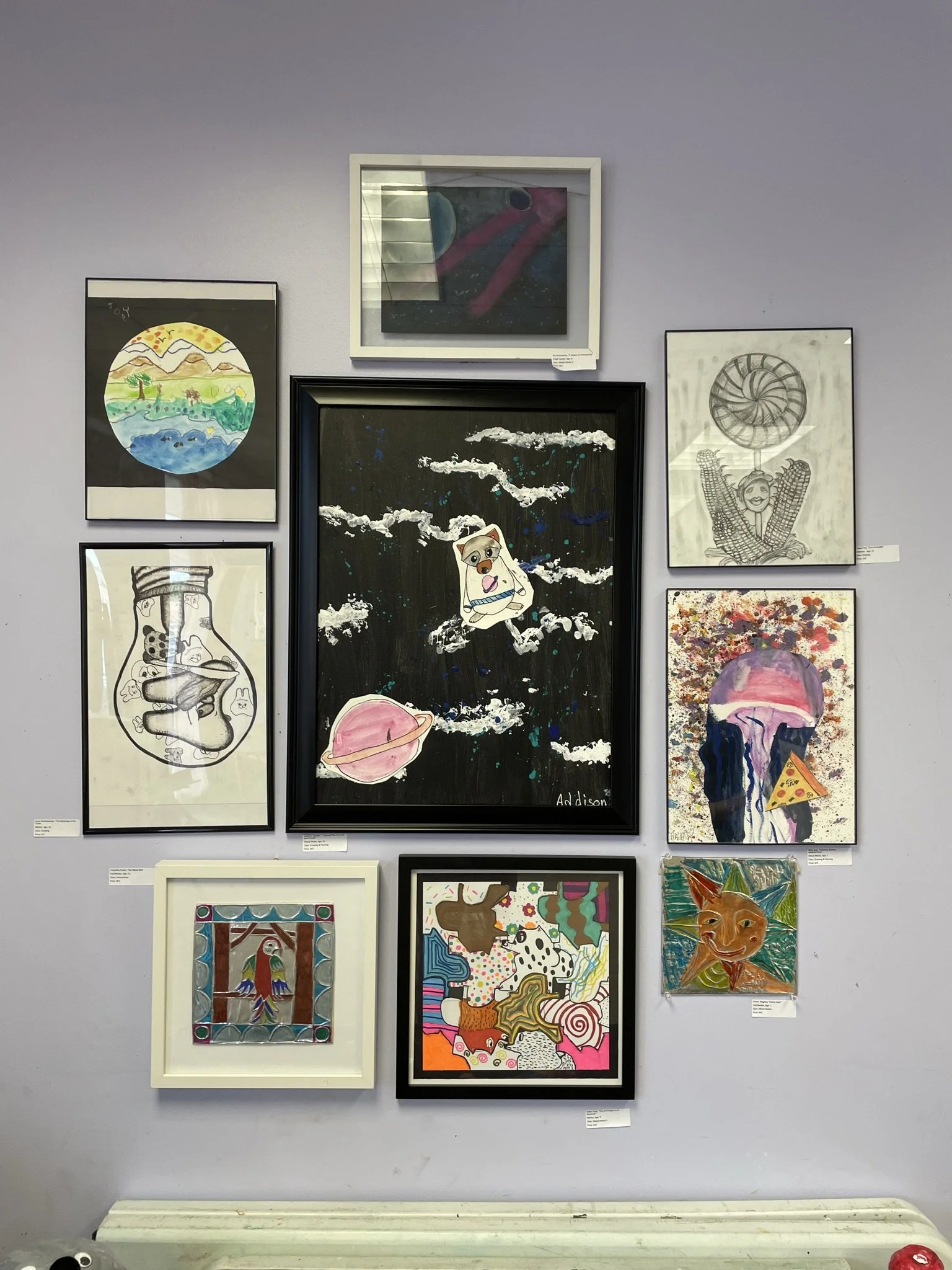 A collection of nine framed artworks on a light purple wall, including drawings of space, sea creatures, abstract patterns, and whimsical characters.