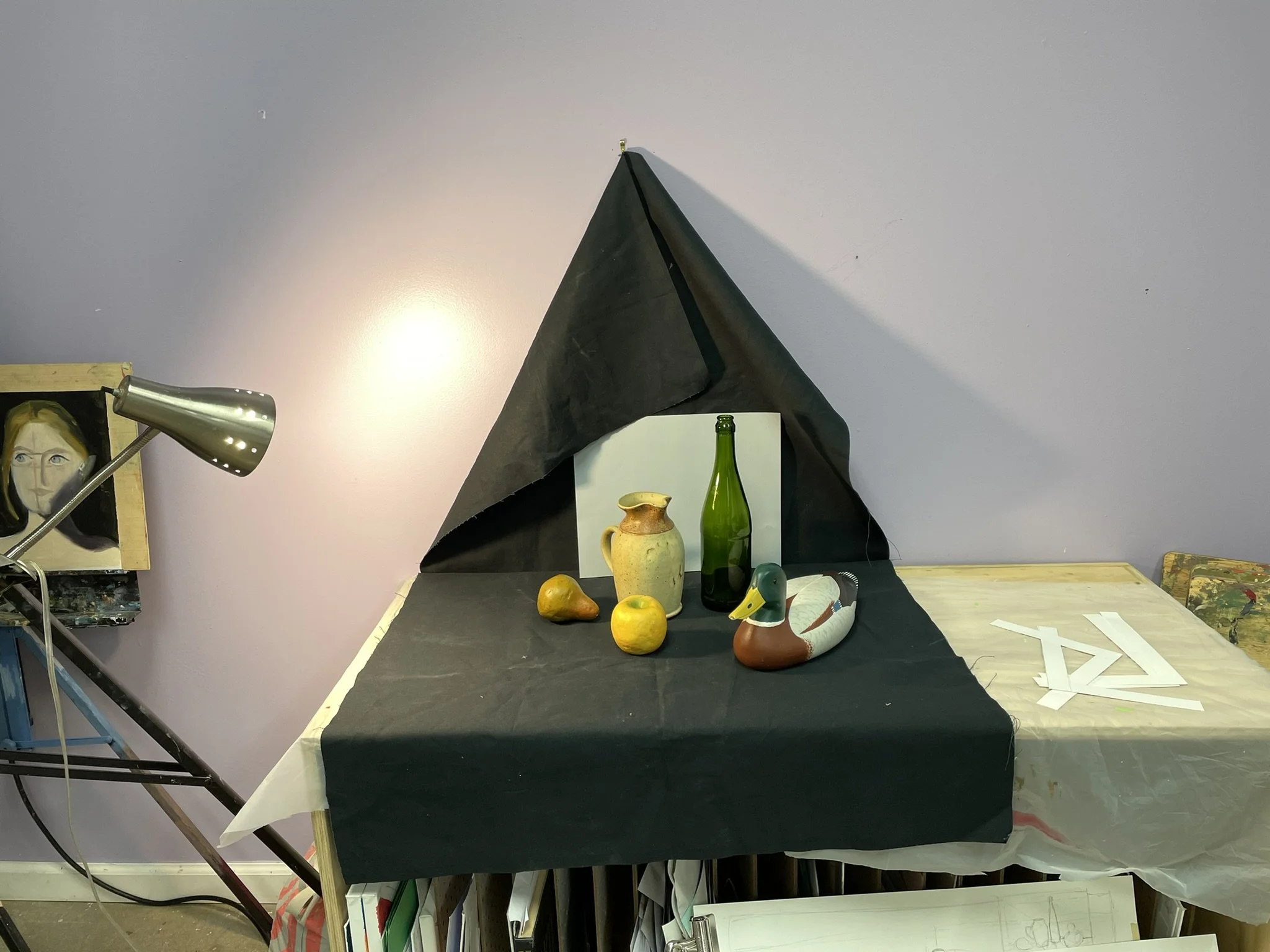 Art studio setup with still life arrangement of a ceramic pitcher, a green bottle, a duck decoy, and two apples on a table draped with black fabric. A black triangular backdrop partly covers a white panel, and a large desk lamp illuminates the scene.