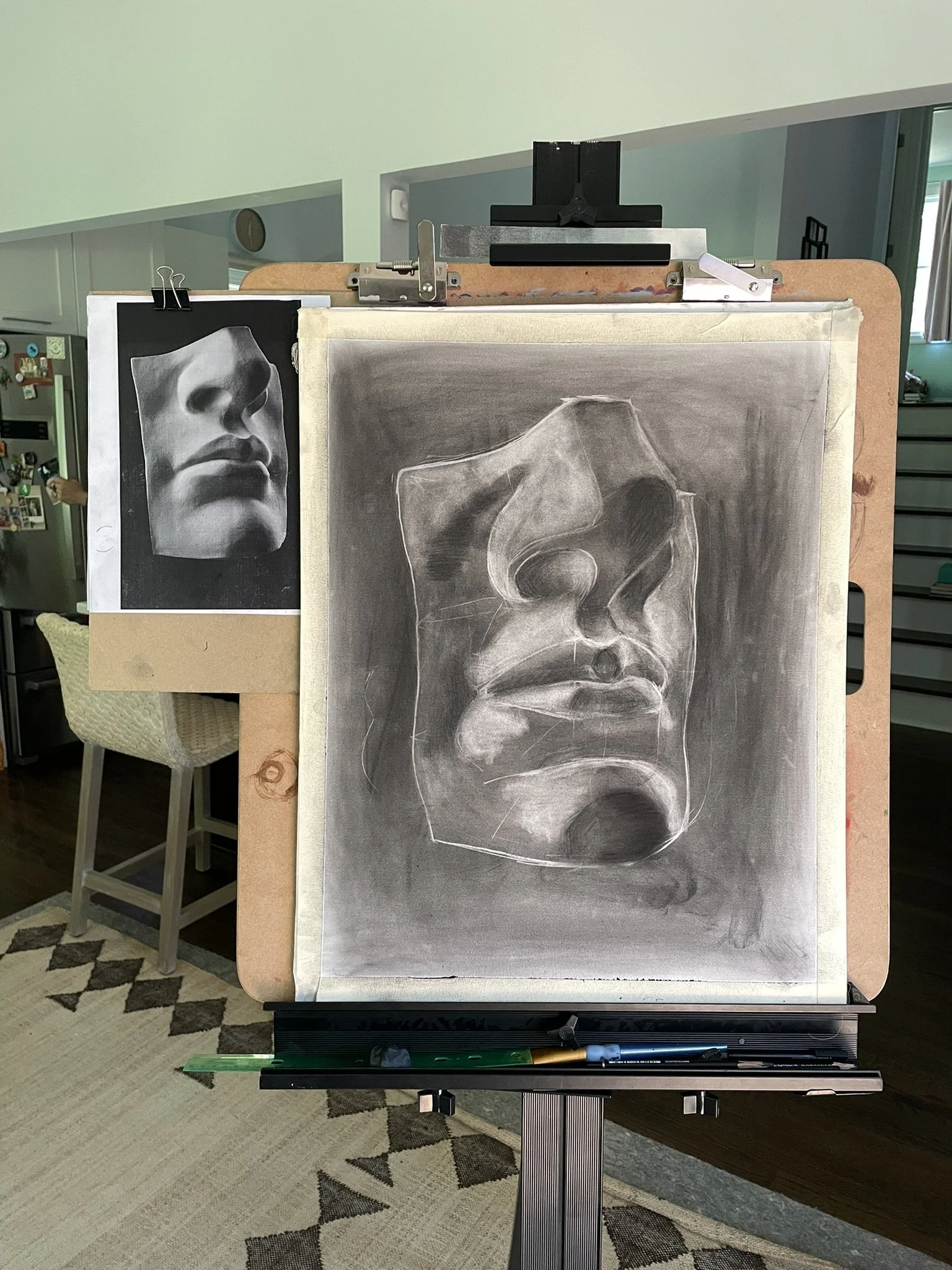 A drawing of a sculpture of a face with one eye missing, in progress on a drawing board, with a reference photo of a woman's face mounted on the side of the board.