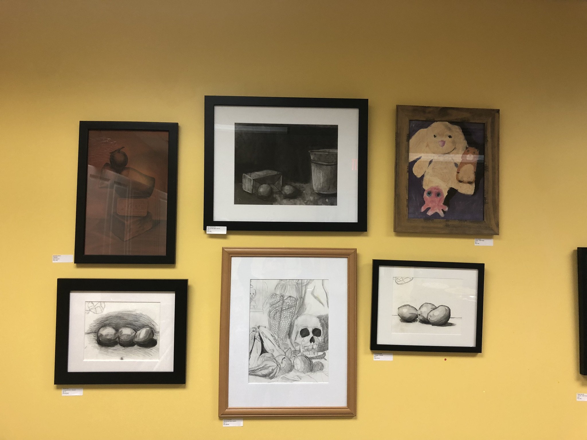 Six framed artworks hanging on a yellow wall in an art gallery.