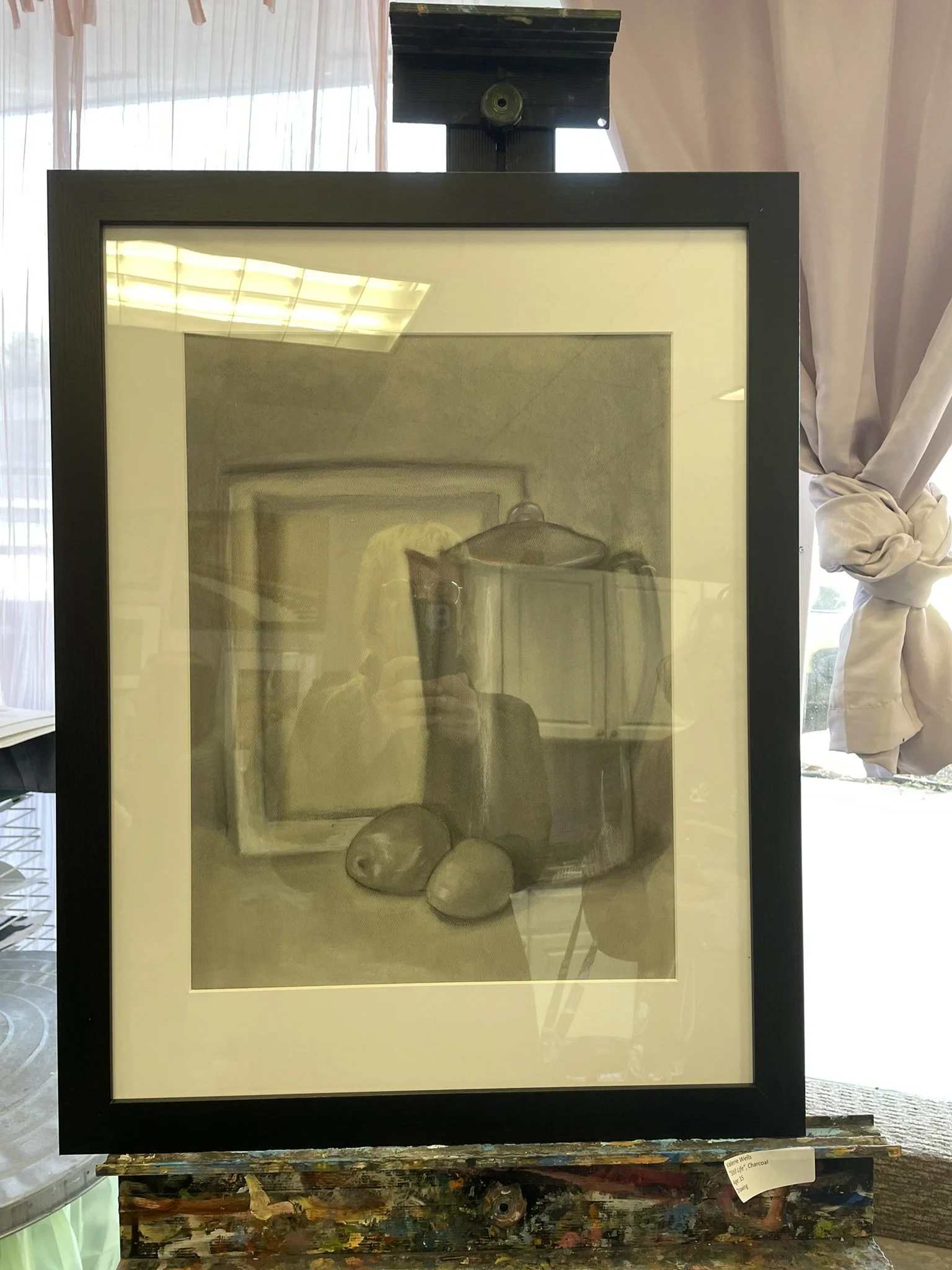 A framed black-and-white still life picture of a jar, scallop shells, and stones on a shelf, displayed in an art gallery with curtains and windows in the background, and someone taking the photo reflected in the glass.