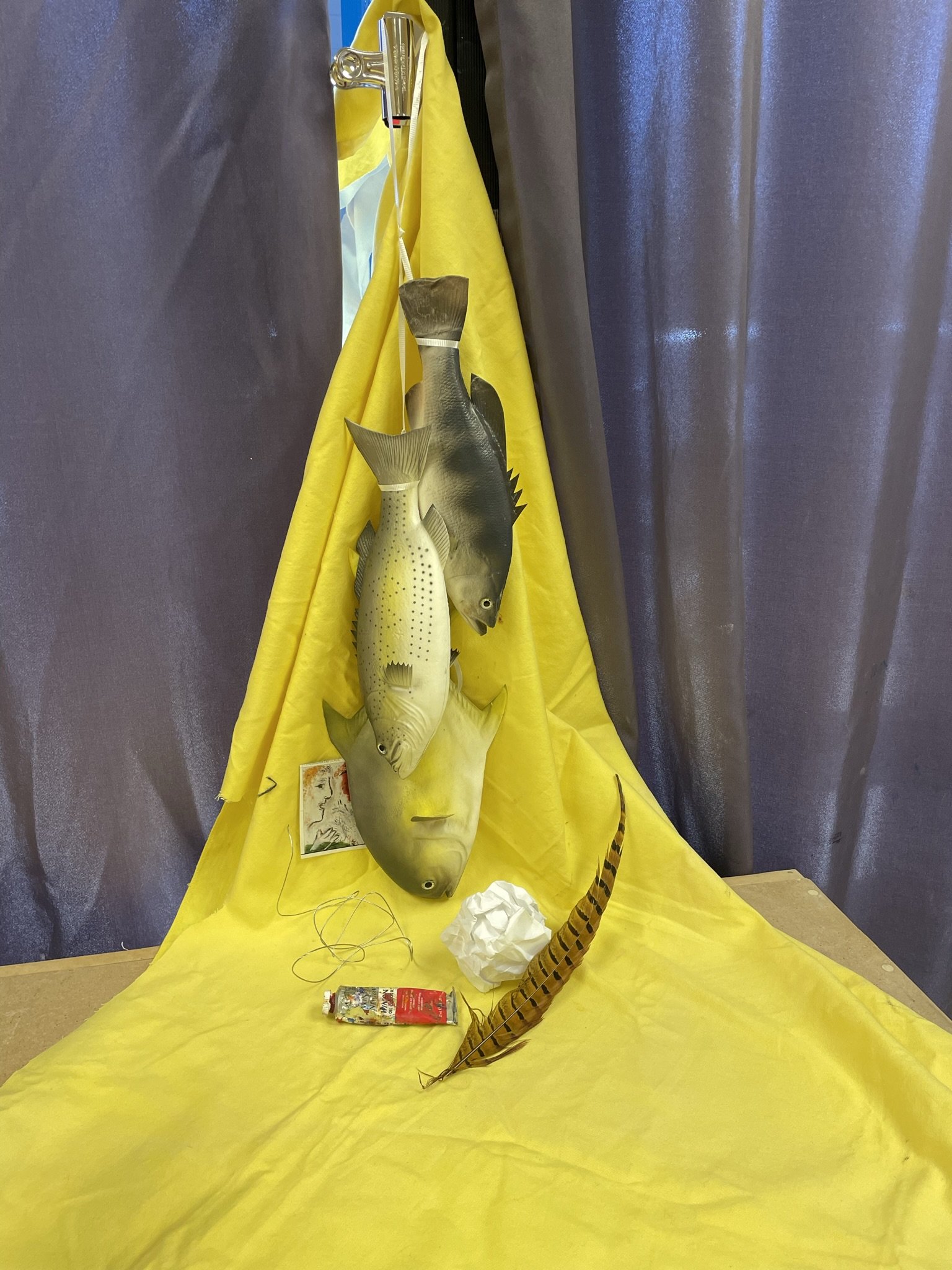 Three realistic fish models hanging on a yellow backdrop, with objects including a feather, crumpled paper, a tube, and a drawing of a woman's face beneath them.