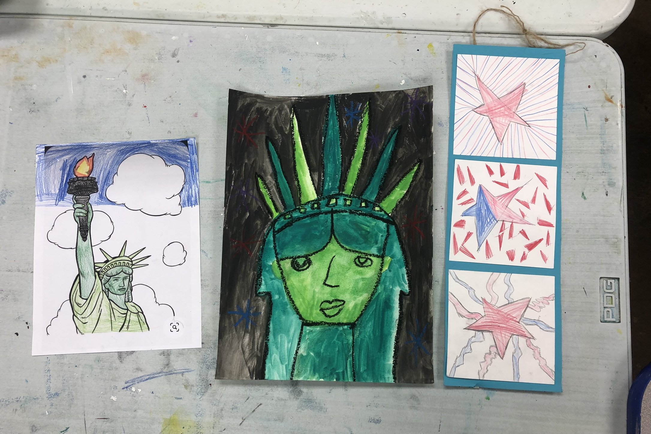 A collection of four art pieces on a table: a coloring page of the Statue of Liberty with clouds, a vibrant drawing of a woman with blue hair and a crown, and a vertical strip of three abstract star-themed artworks with red and blue colors.