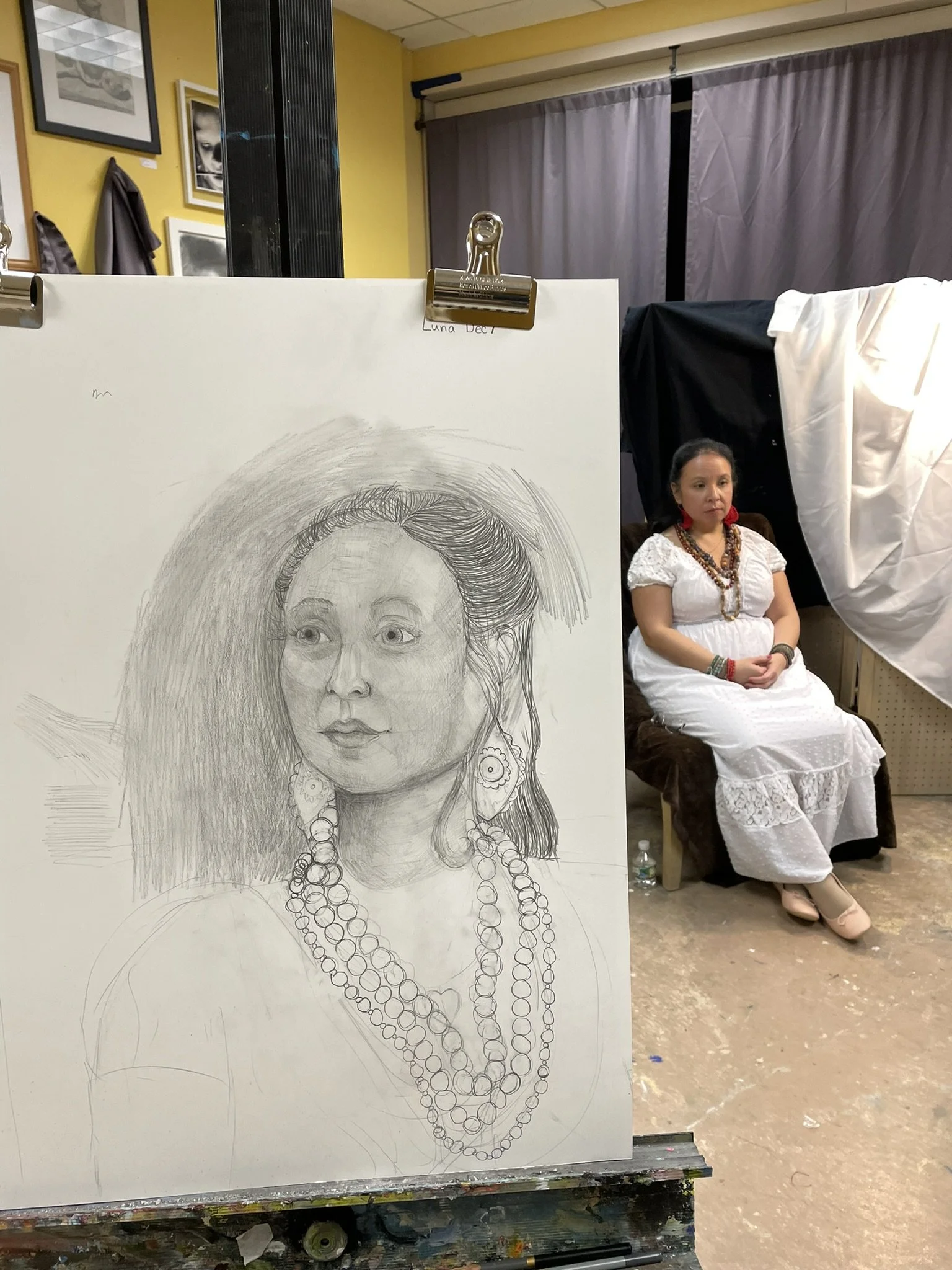 A detailed pencil sketch of a woman wearing large earrings and multiple necklaces, positioned in front of a woman dressed in a white dress, sitting on a chair in an art studio.