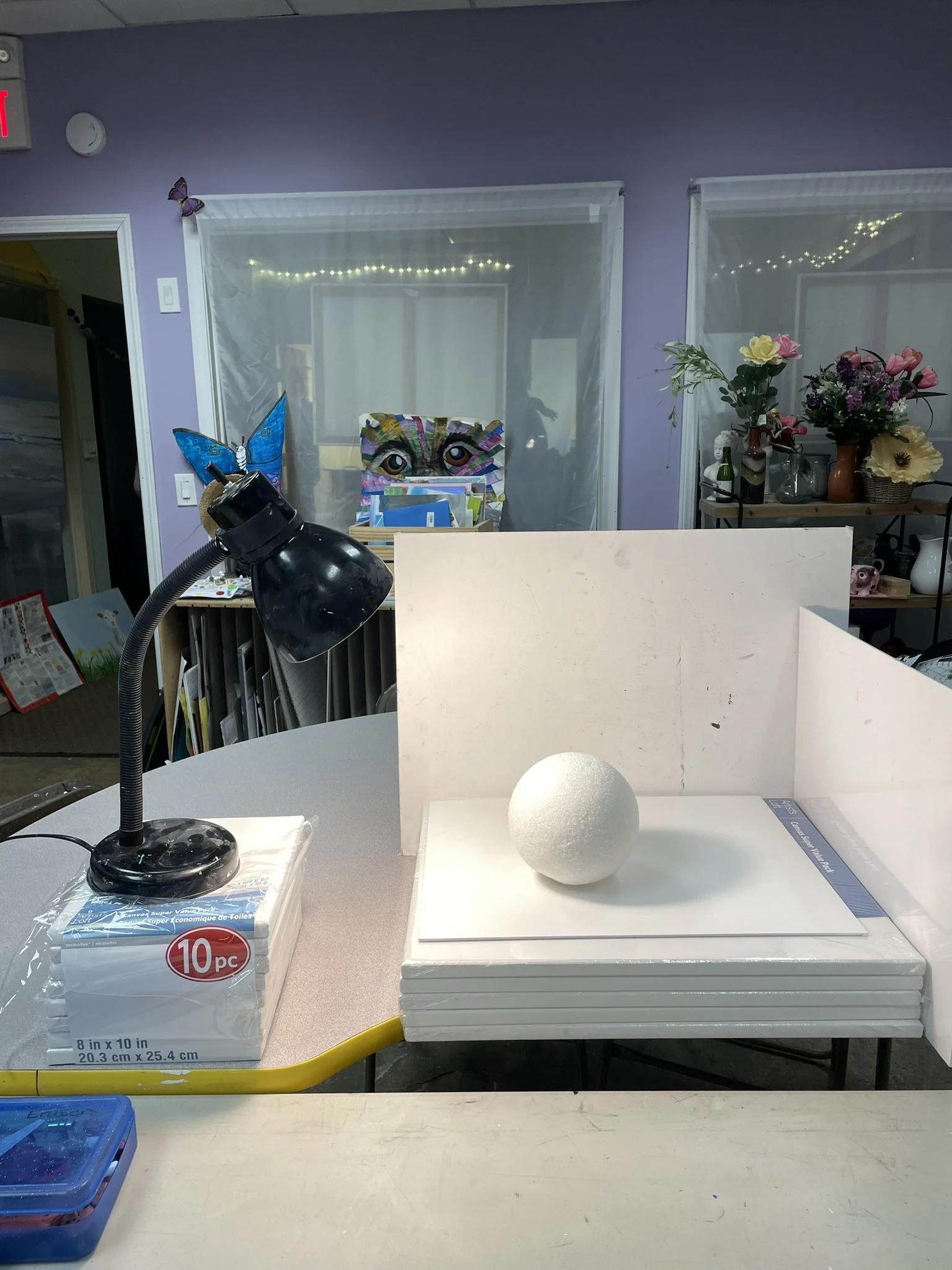 Art studio workspace with a black desk lamp, a stack of white foam boards, a white foam sphere, and a partition, with art supplies and colorful artwork and decorations in the background.