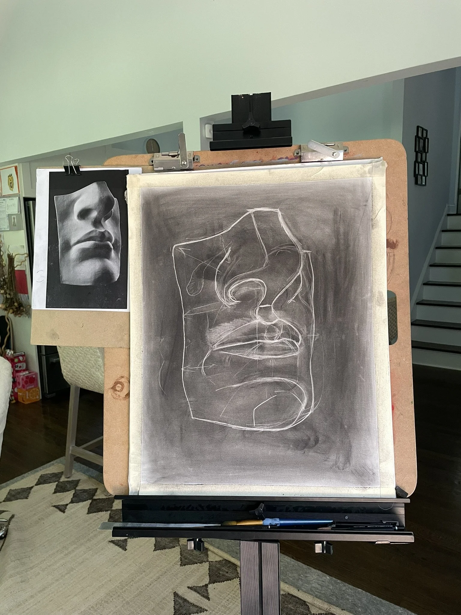 An art easel displays a charcoal sketch of a human face viewed from below, with a reference photo of the same face on the side.