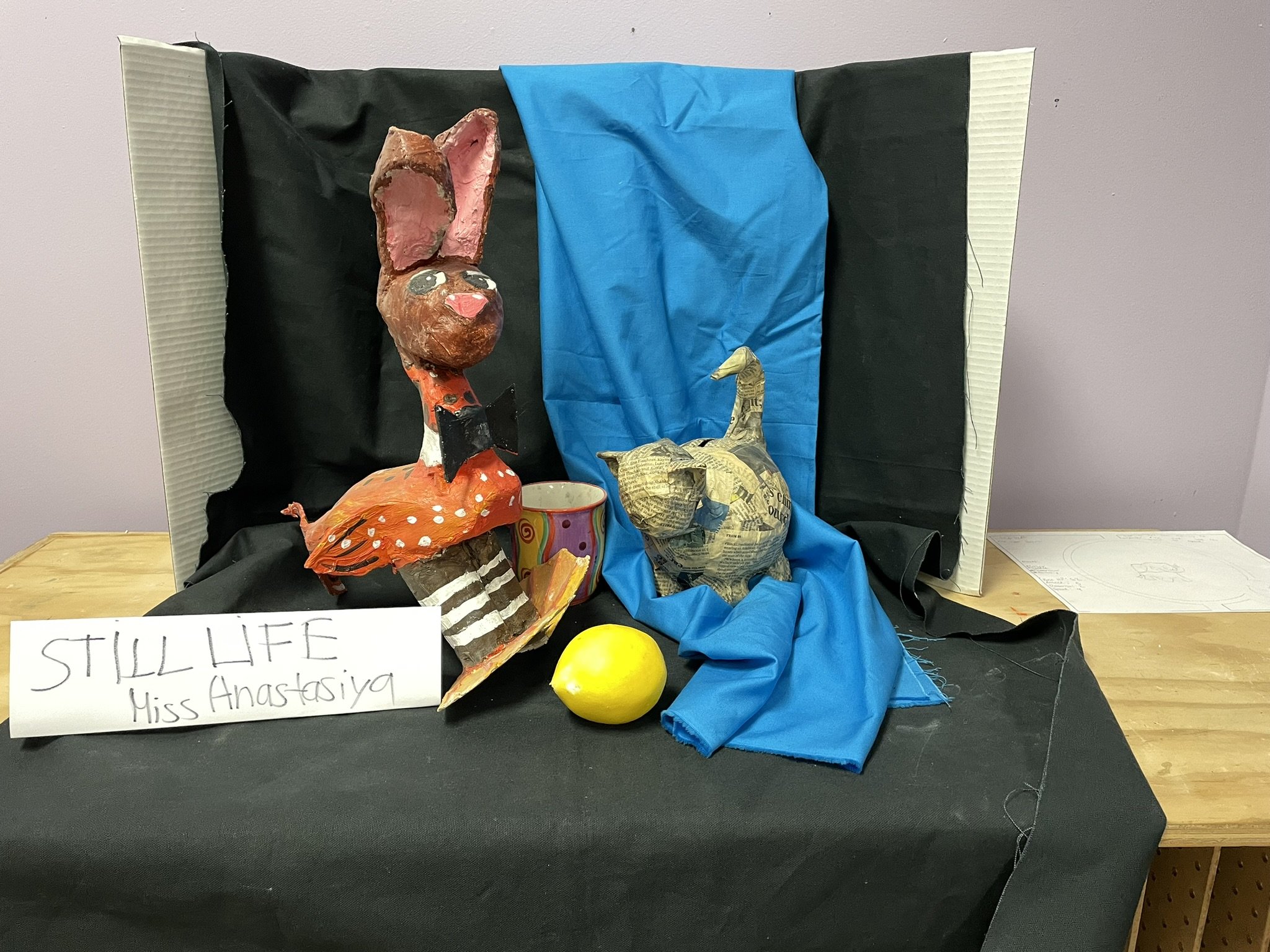 A diorama depicting a colorful clay or paper-mâché rabbit, a duck made from newspaper, a yellow lemon, and a marbled cup, with a black and blue backdrop, and a sign reading 'STILL LIFE Miss Anastasiya' displayed in front.