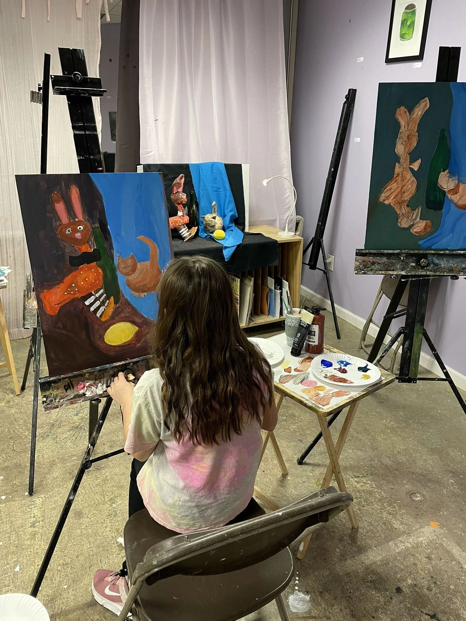 A girl with long wavy hair painting a colorful still life scene on a canvas in an art studio. The scene features a bunny plush toy, an orange cat, books, a yellow oval, and a plate, rendered in a playful style.