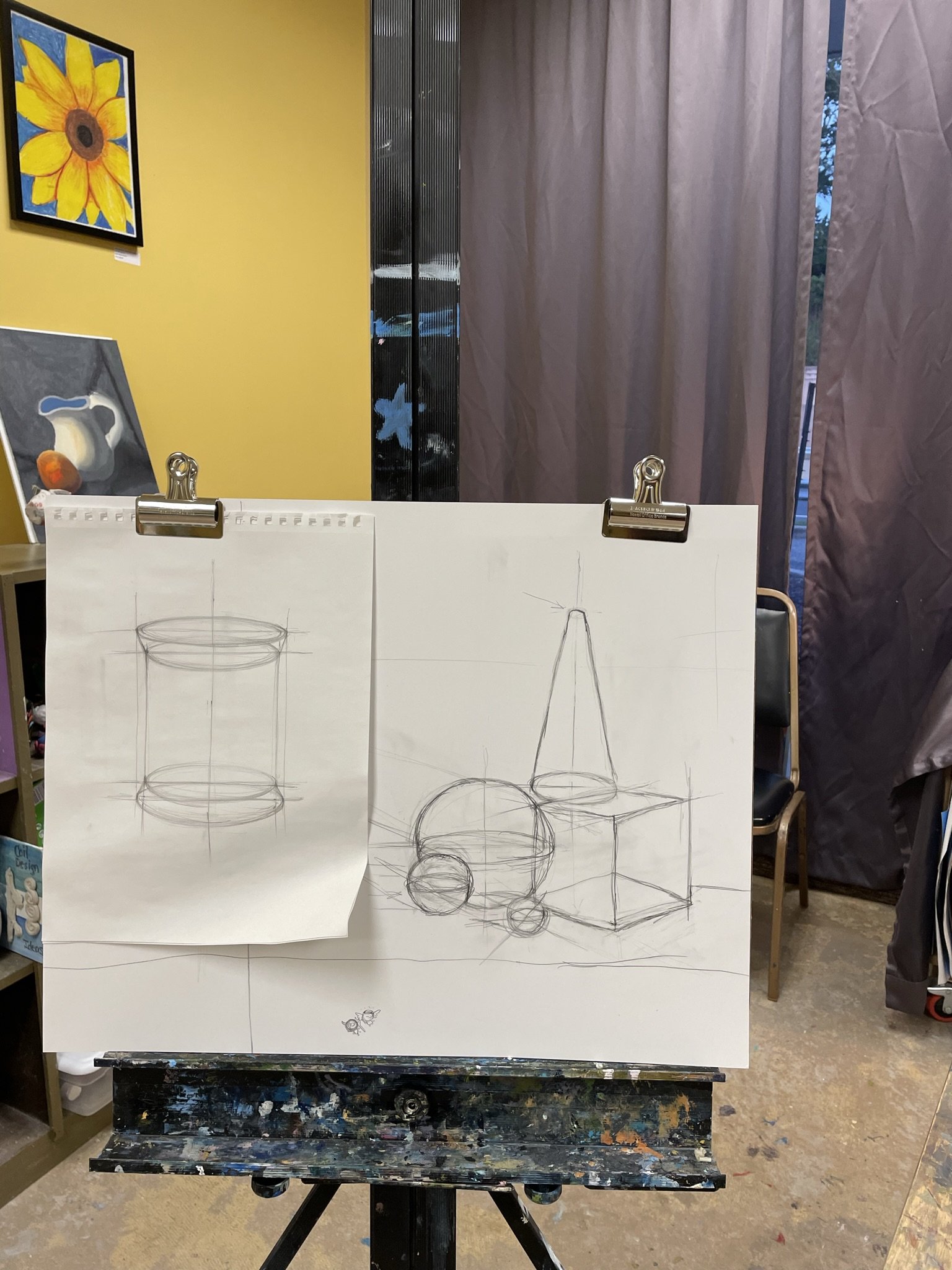 Sketches of geometric shapes, including a cylinder, cone, sphere, and cube, on a large sheet of paper clipped to an easel in an art studio.