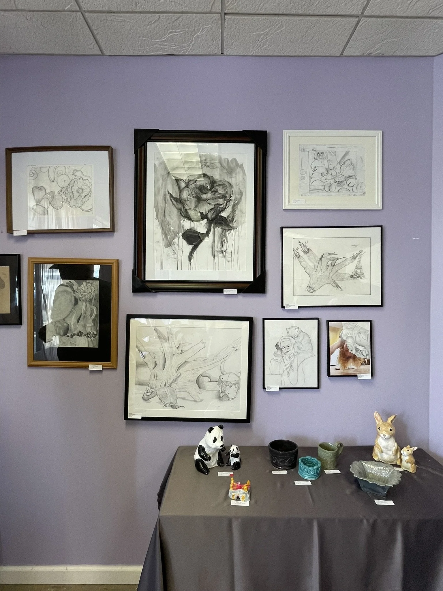 An art display featuring framed sketches hung on a light purple wall and three ceramic animal figurines on a table below. The sketches include various subjects such as flowers, animals, and human figures.