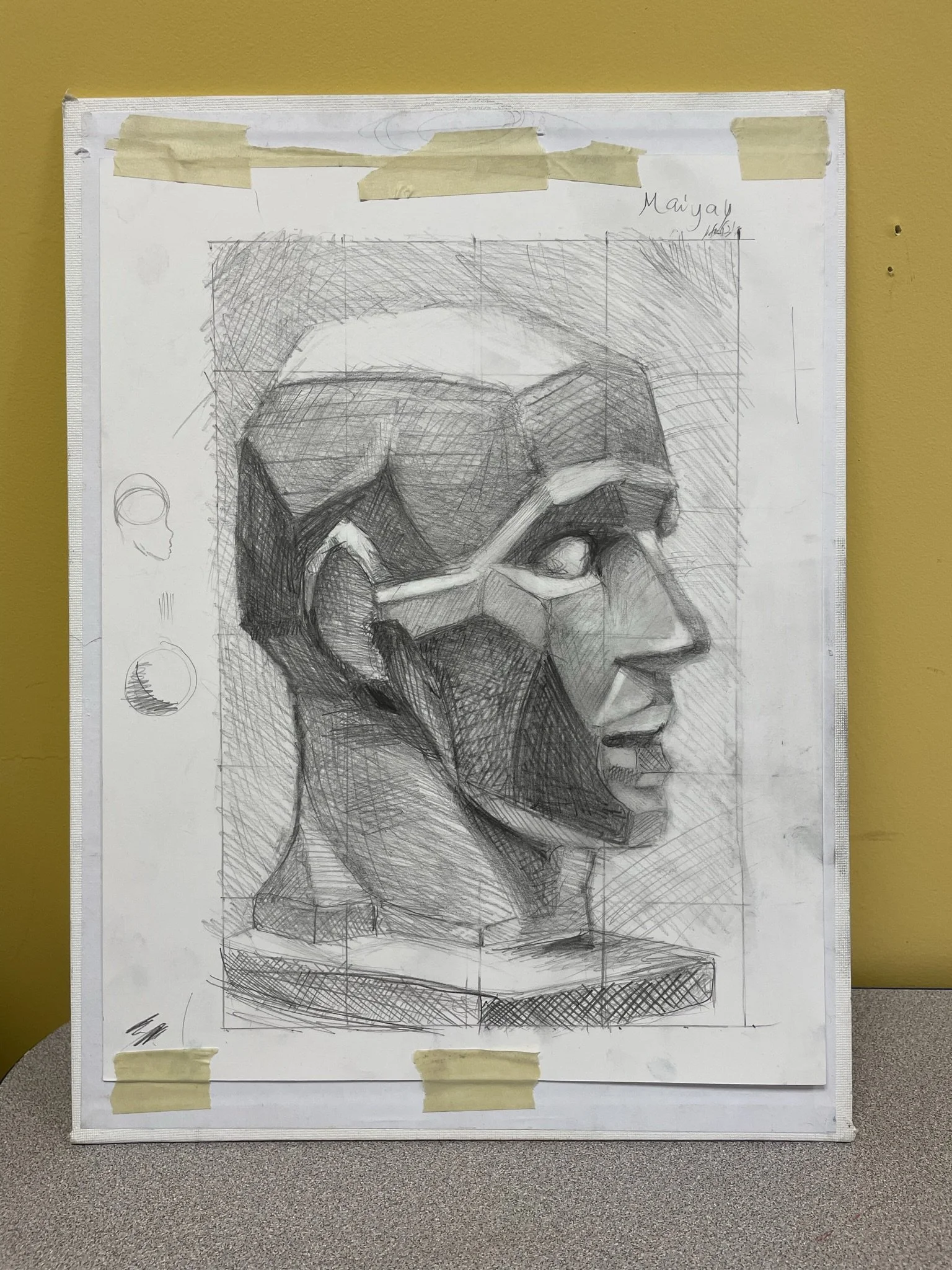 A detailed graphite sketch of a human head in profile, with geometric and angular features, wearing glasses, mounted on a board taped at the top and bottom, with the name 'Maiyal' and date '16/5/23' written in the top right corner.