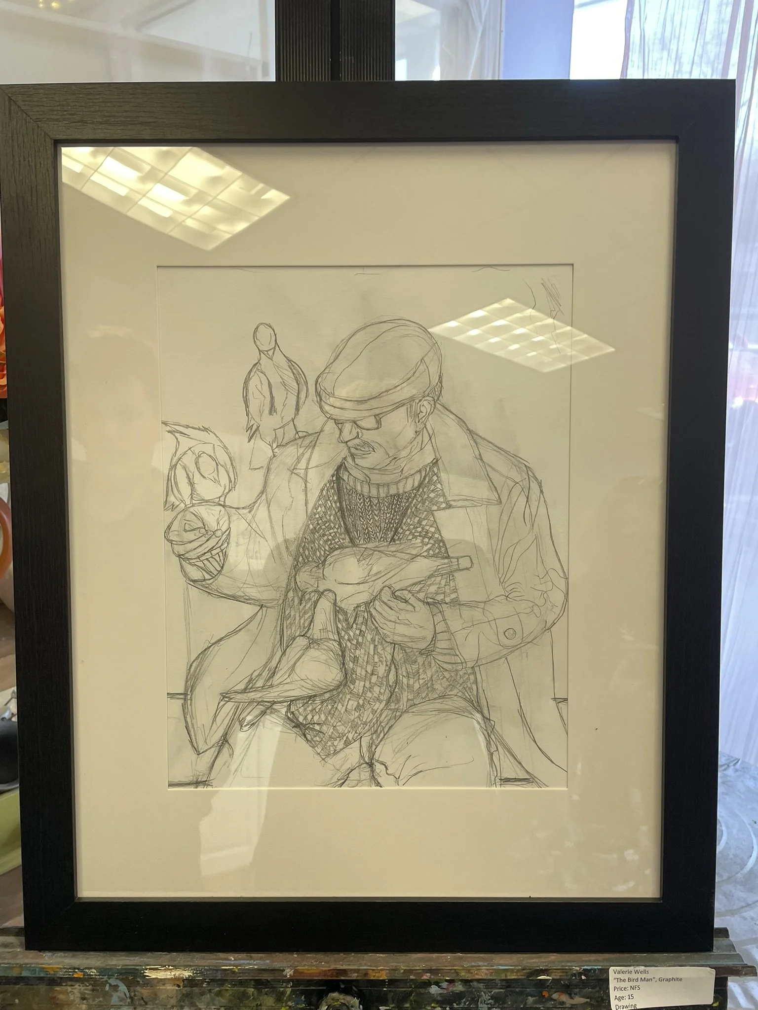 A framed sketch of a man in a hat and glasses holding a shoe, with two women standing behind him, one on his left and one in the background to his right.