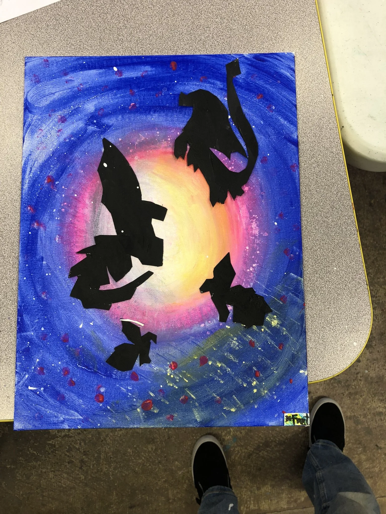 Painted canvas of a galaxy with a yellow and pink center, surrounded by blue and purple. Black paper cutouts of bats arranged in a circular pattern overlay the galaxy.