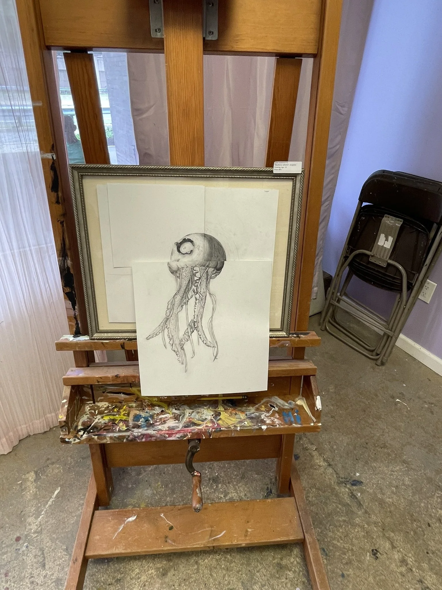 A framed drawing of a jellyfish leaning against a woodworking easel with paint stains, in an art studio. Behind, there are stacked folding chairs and part of a purple wall.