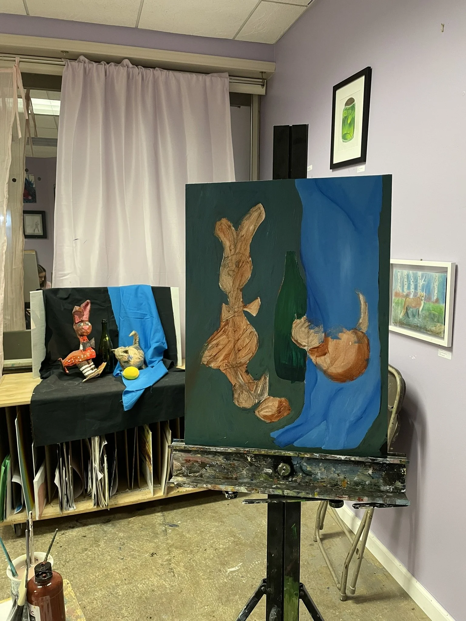 An art studio with a painting on an easel, depicting a still life scene with a rabbit-shaped wooden sculpture, a green bottle, and a round brown object against a blue background. There are also other small sculptures or objects on a table covered wit