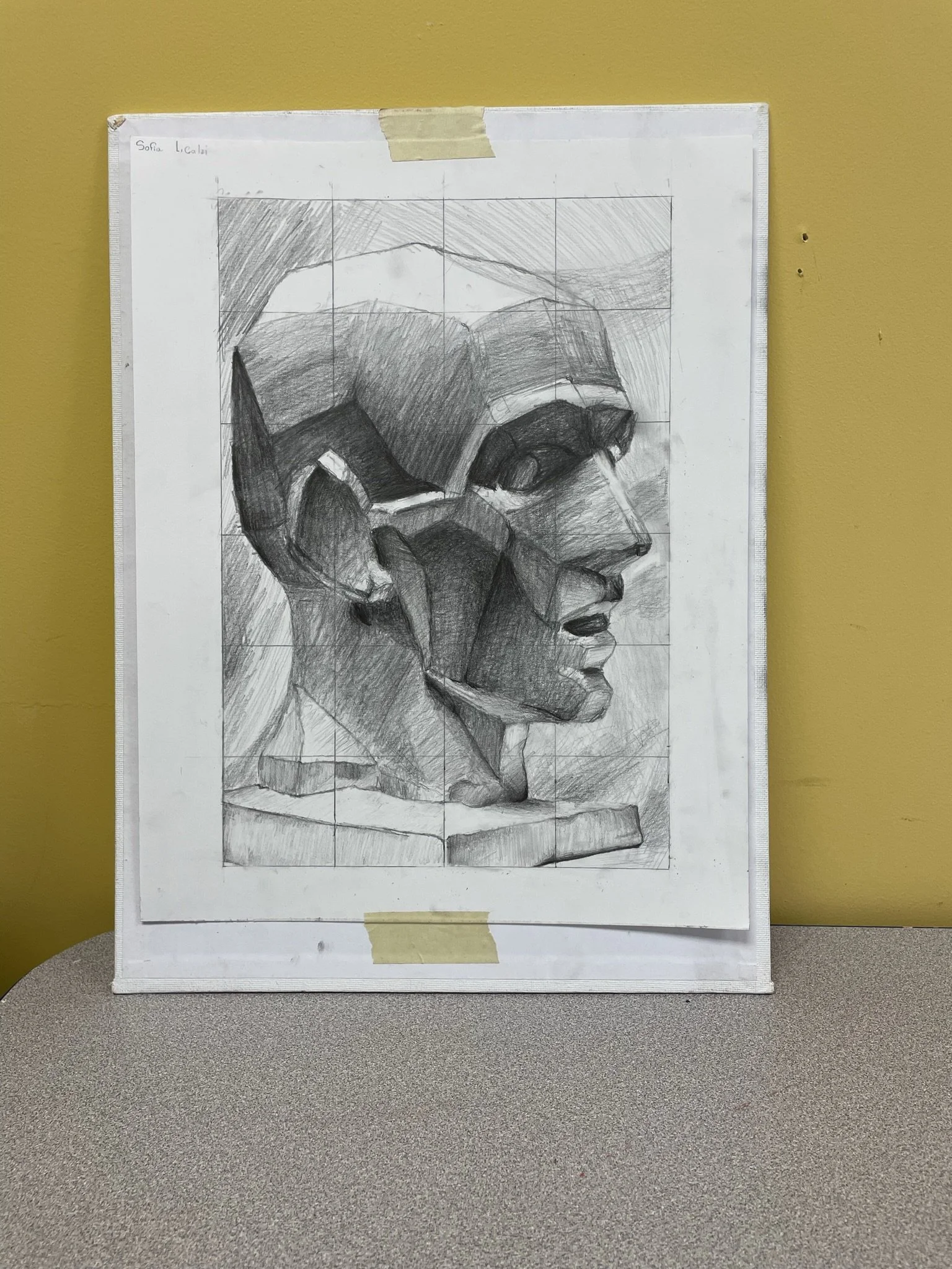 A pencil drawing on grid paper of a Cubist-style profile of a man's face, taped to a yellow wall.
