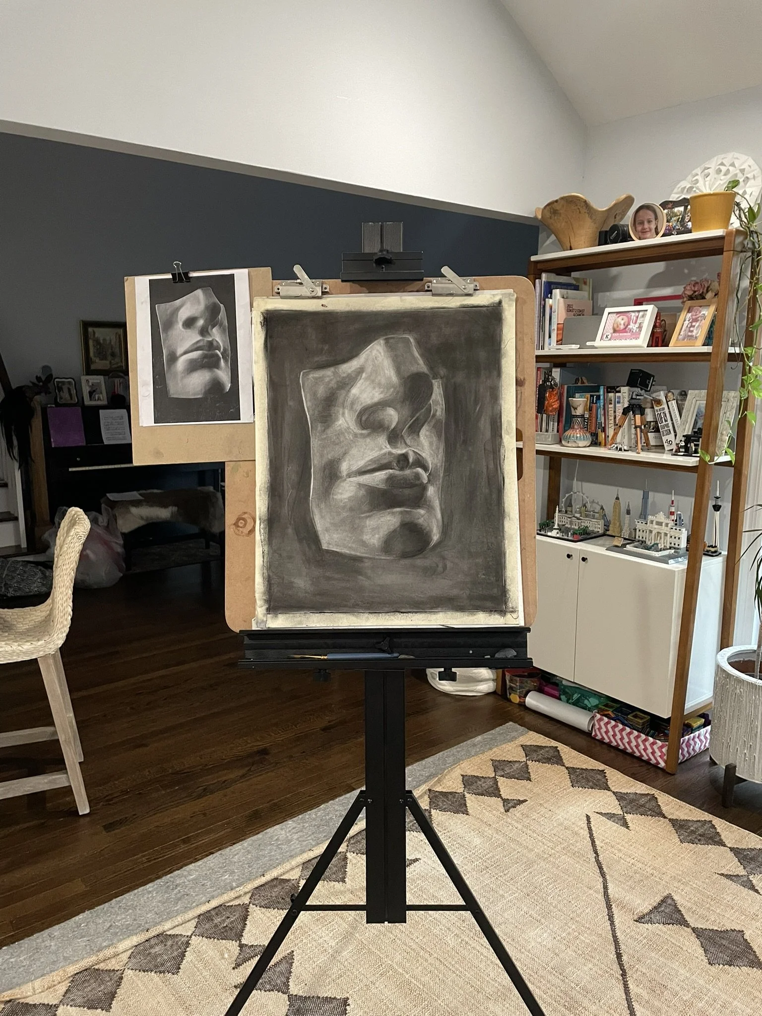 A charcoal drawing of a human face in a three-quarter view on an easel, with a reference photo of the same face pinned to the side. The room has a bookshelf with decorative items and framed photos, a woven chair, and a patterned rug.
