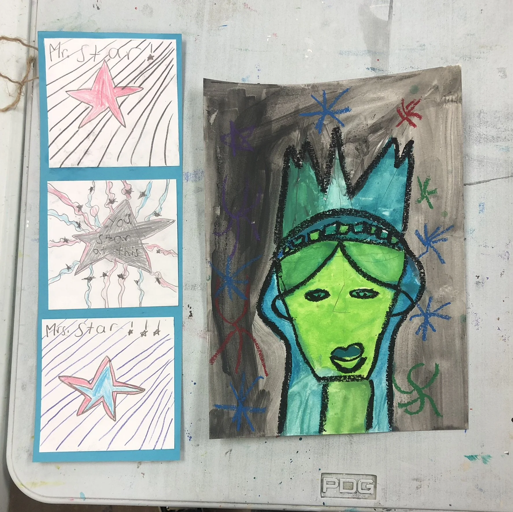 Colorful drawing of a green face with black outlines, wearing a crown, against a black background with blue, red, purple, and green star-shaped scribbles. Three smaller colored star drawings on the left include the words 'Mr. Star' and 'Star of This'