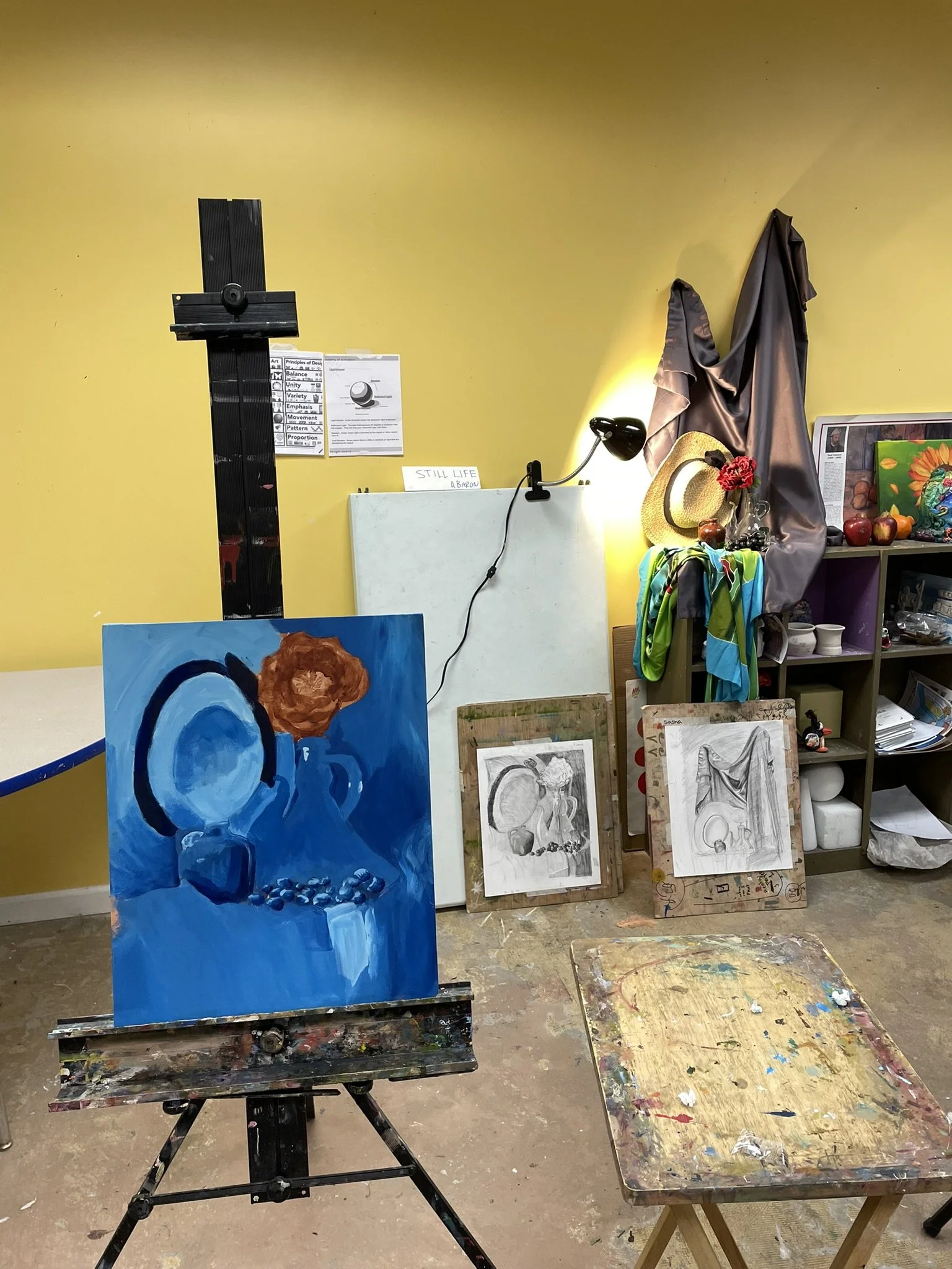 An art studio with an easel holding a blue painting of a teapot, a still life setup with a straw hat, flowers, and apples, and several sketches and paintings on the floor and shelves. The studio has a yellow wall and a nearby table with paint supplie