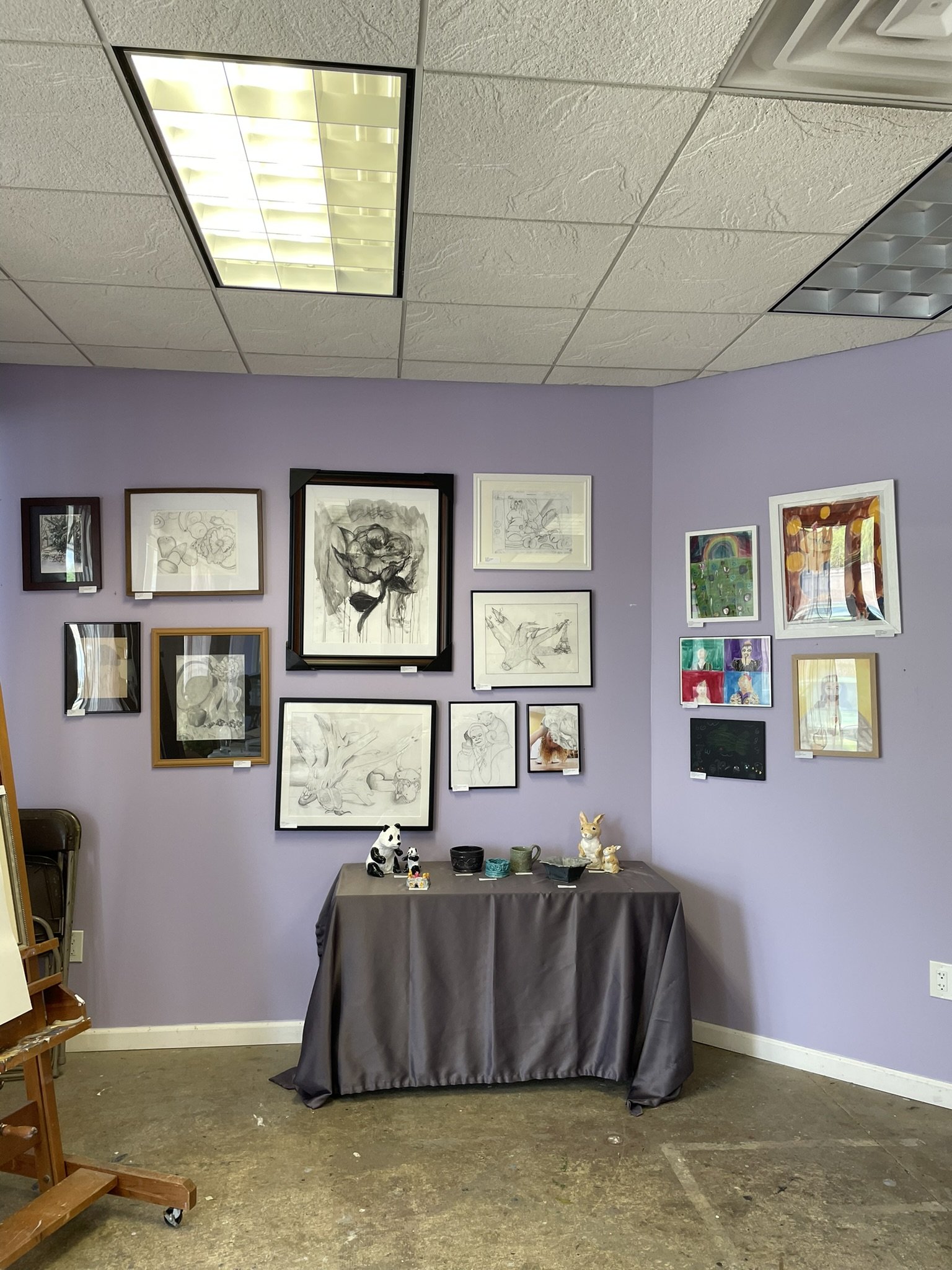 An art gallery corner with framed artwork on lavender walls, a black table with various ceramic figurines, and ceiling fluorescent lights.