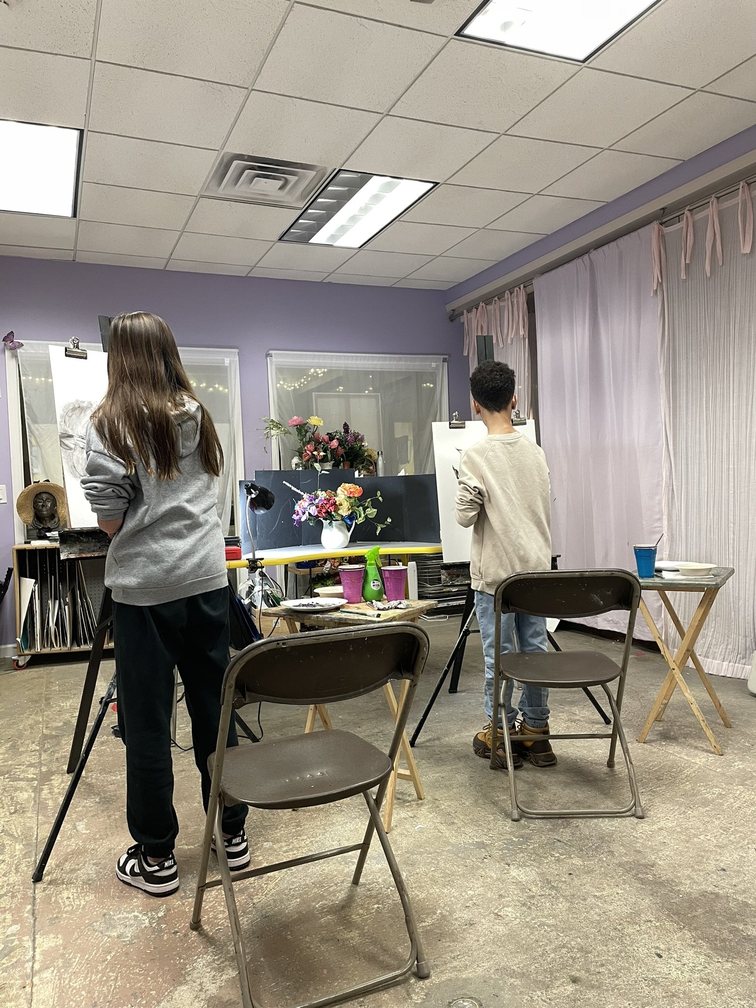 Two people drawing or painting in a classroom or art studio with tables, chairs, flowers, and art supplies.