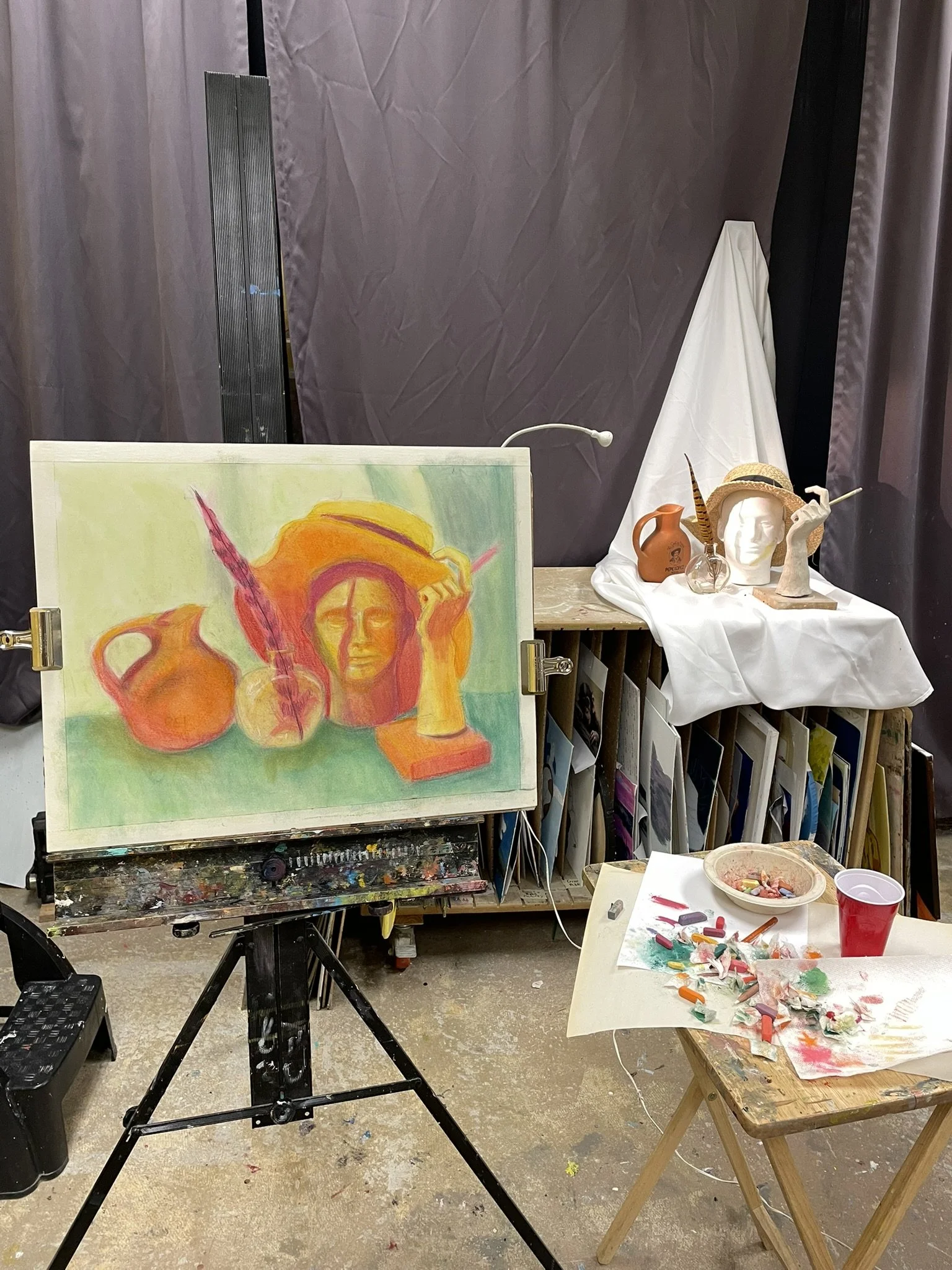 An art studio setup with a colorful painting on an easel, featuring a woman's face, vases, and a feather. In the background, a display table holds a clay bust, a vase, and a straw hat. Art supplies, paint, and brushes are spread on a small wooden tab