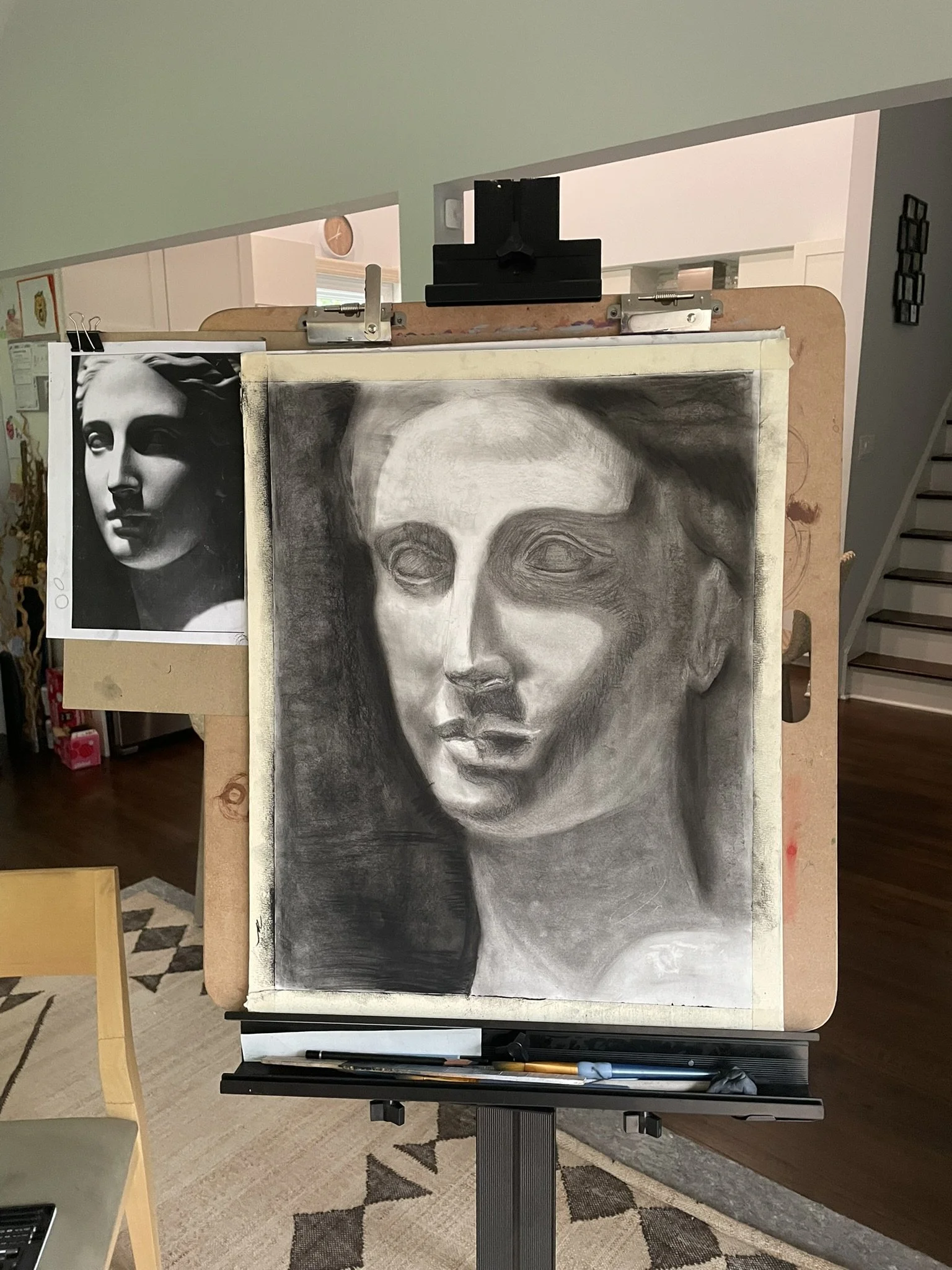 Art easel with a black and white portrait drawing of a woman's face, partially completed, with a printed reference image clipped on the side.