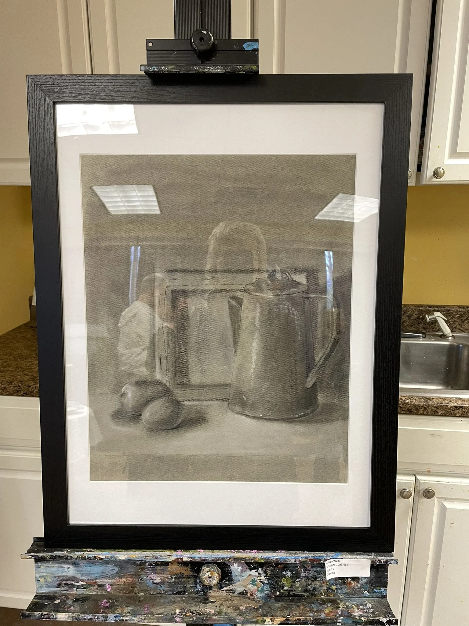 A grayscale still life painting of a teapot, two eggs, and a box, with a framed mirror hanging above, reflected in the glass.