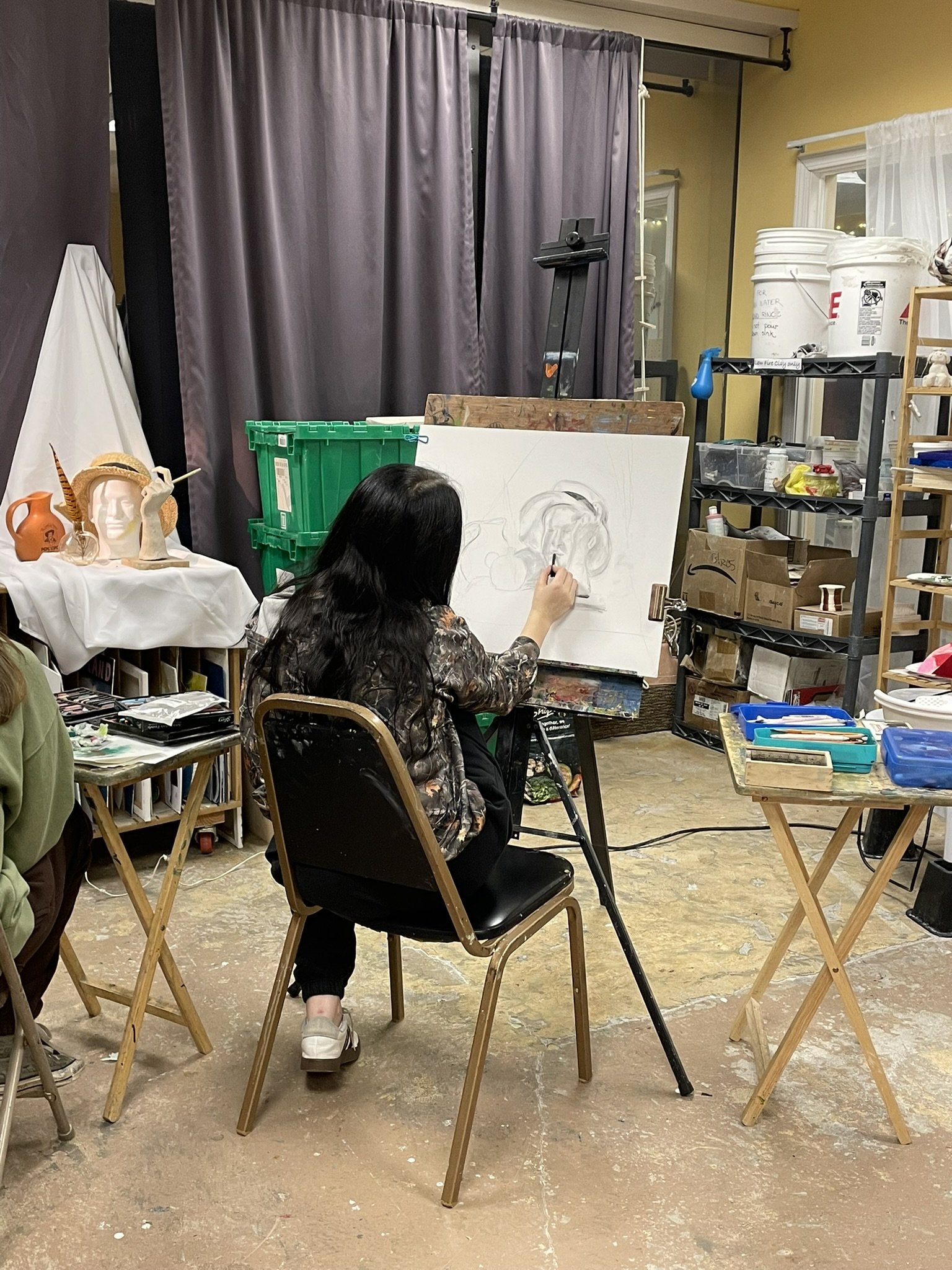 A woman with long black hair is sitting on a black chair, drawing on a large white canvas set up on an easel in an art studio. The studio has various art supplies, shelves, and decorations around. There are dark grey curtains and white walls, and a s