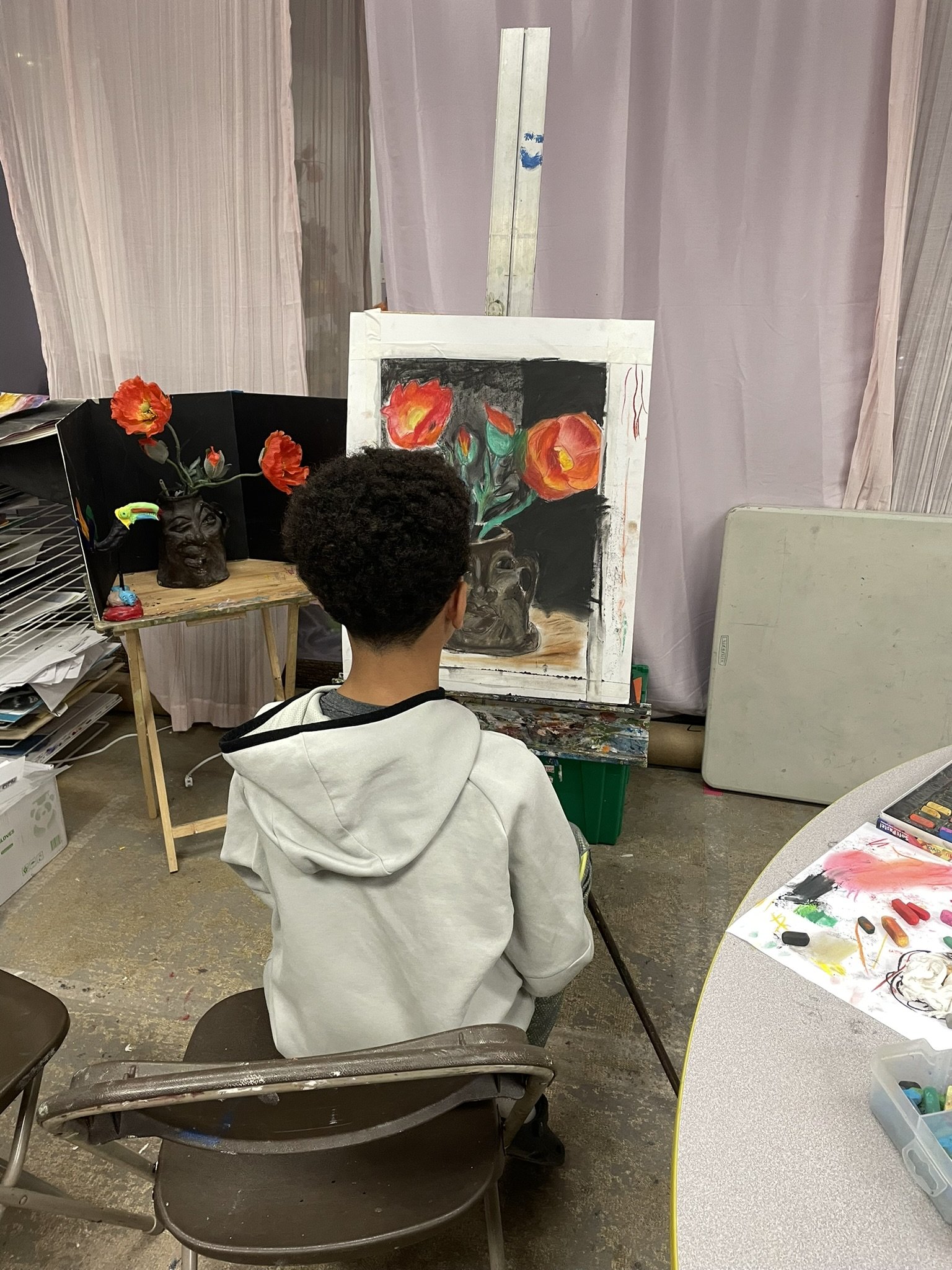 A person with short curly hair in a gray hoodie, viewed from behind, sitting and painting a still life with orange and red flowers in a black vase on a white canvas in an art studio.