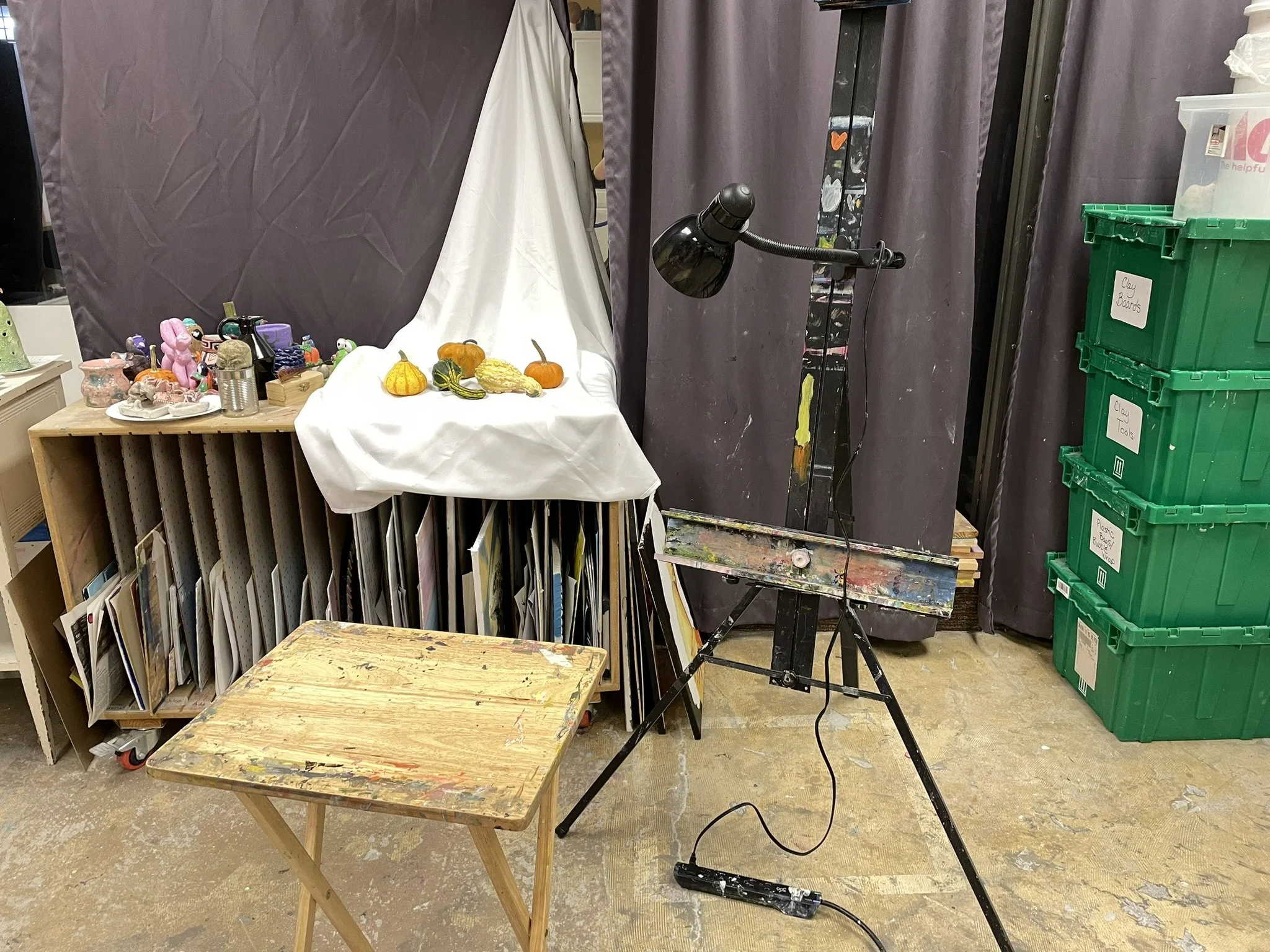 An art studio setup with a wooden table, a lampshade on an easel, a shelf with various objects, and a white cloth displaying small gourds and pumpkins. There are green storage bins labeled 'Clay Boards', 'Clay Tools', and 'Plaster Casts' on the right