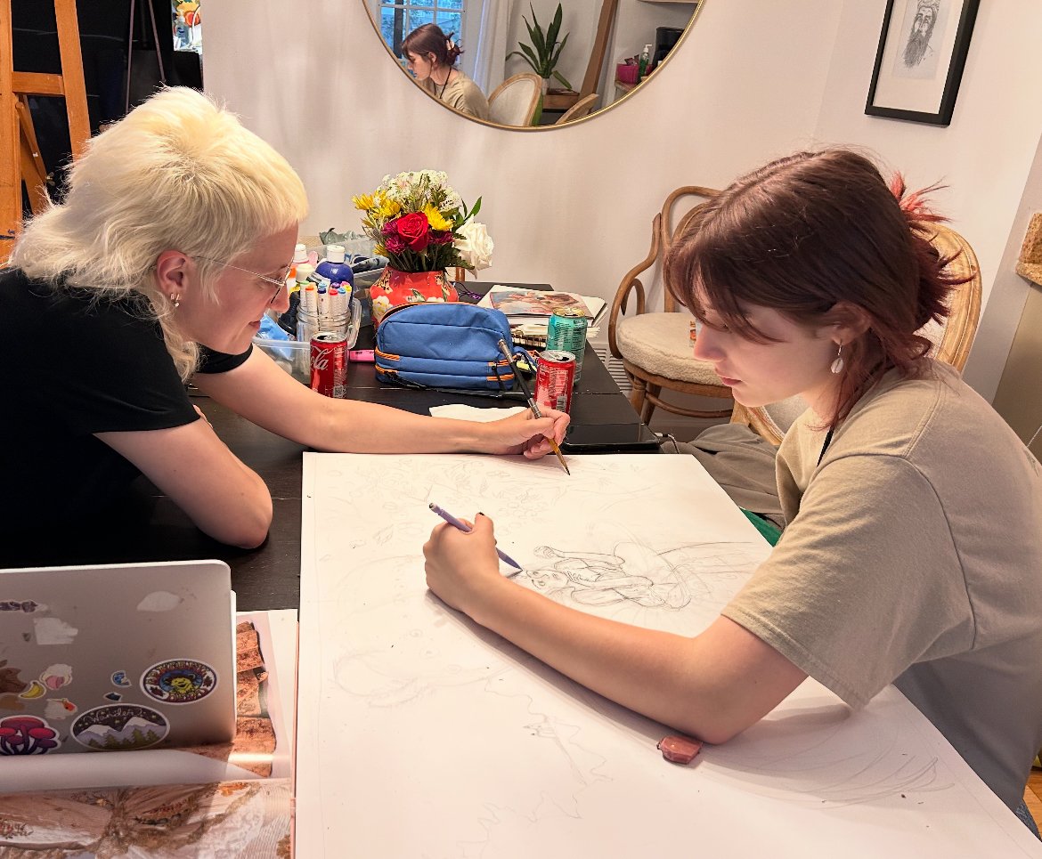 Two women are drawing on a large sheet of paper at a table. One woman with blonde hair is sketching while the other woman with brown hair focuses on her work. The table has various art supplies, cans, and a bouquet of flowers. A mirror and a framed p