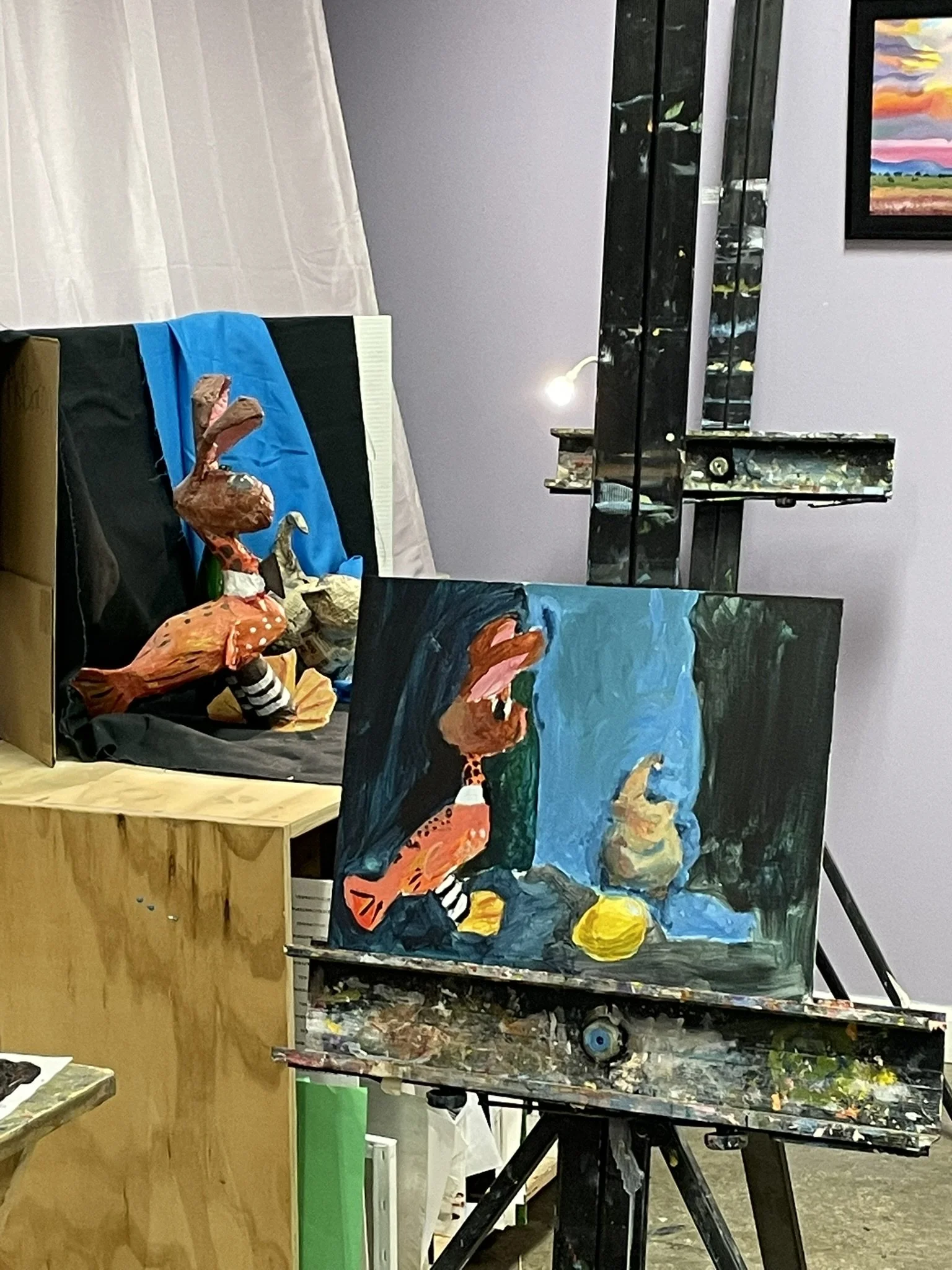 Paintings of a dog with a long neck, with one showing the dog sitting and the other showing the dog with its mouth open, on an artist's easel in an art studio.