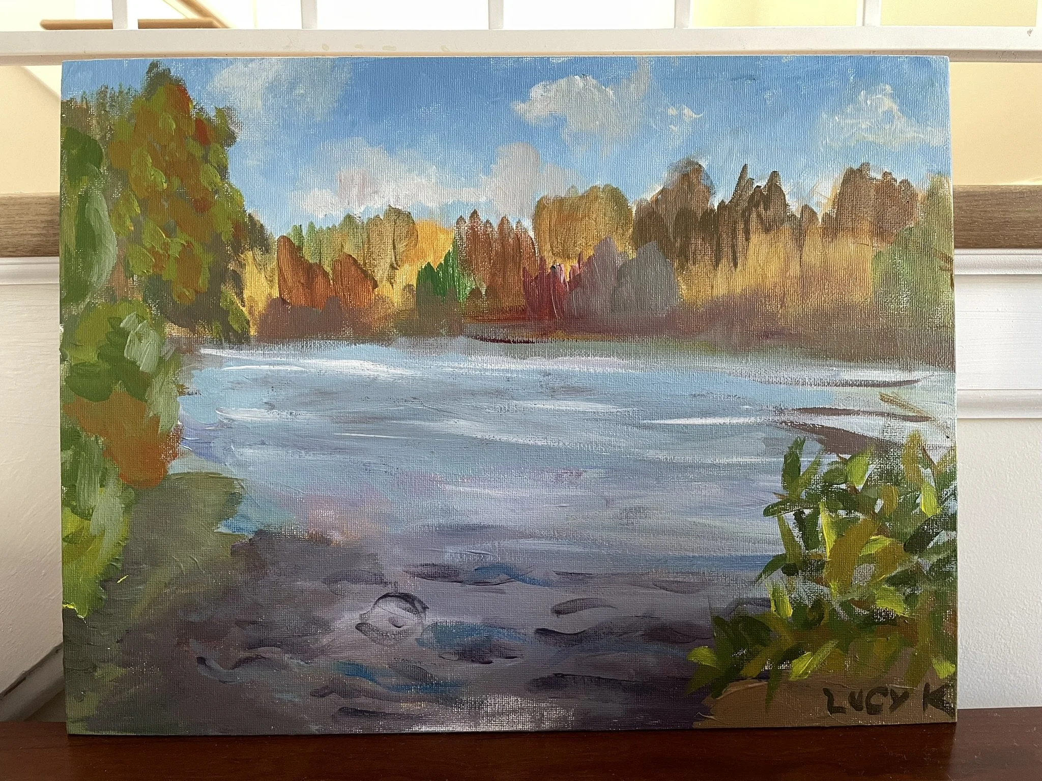 An oil painting of a river scene surrounded by trees with green, orange, and yellow foliage under a partly cloudy blue sky.