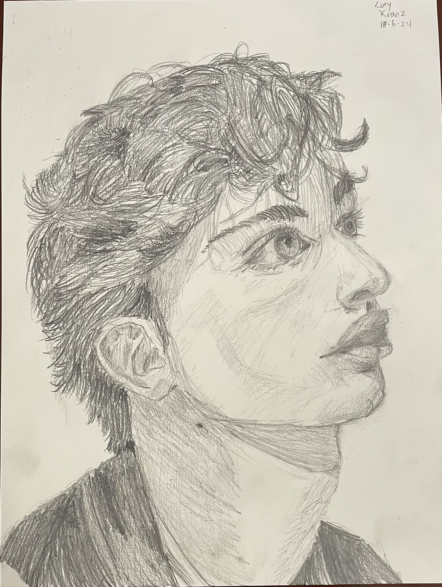 A detailed pencil sketch of a person's profile with short, wavy hair, prominent eye, nose, and lips, signed by Lucy Kranz and dated 10-6-24.