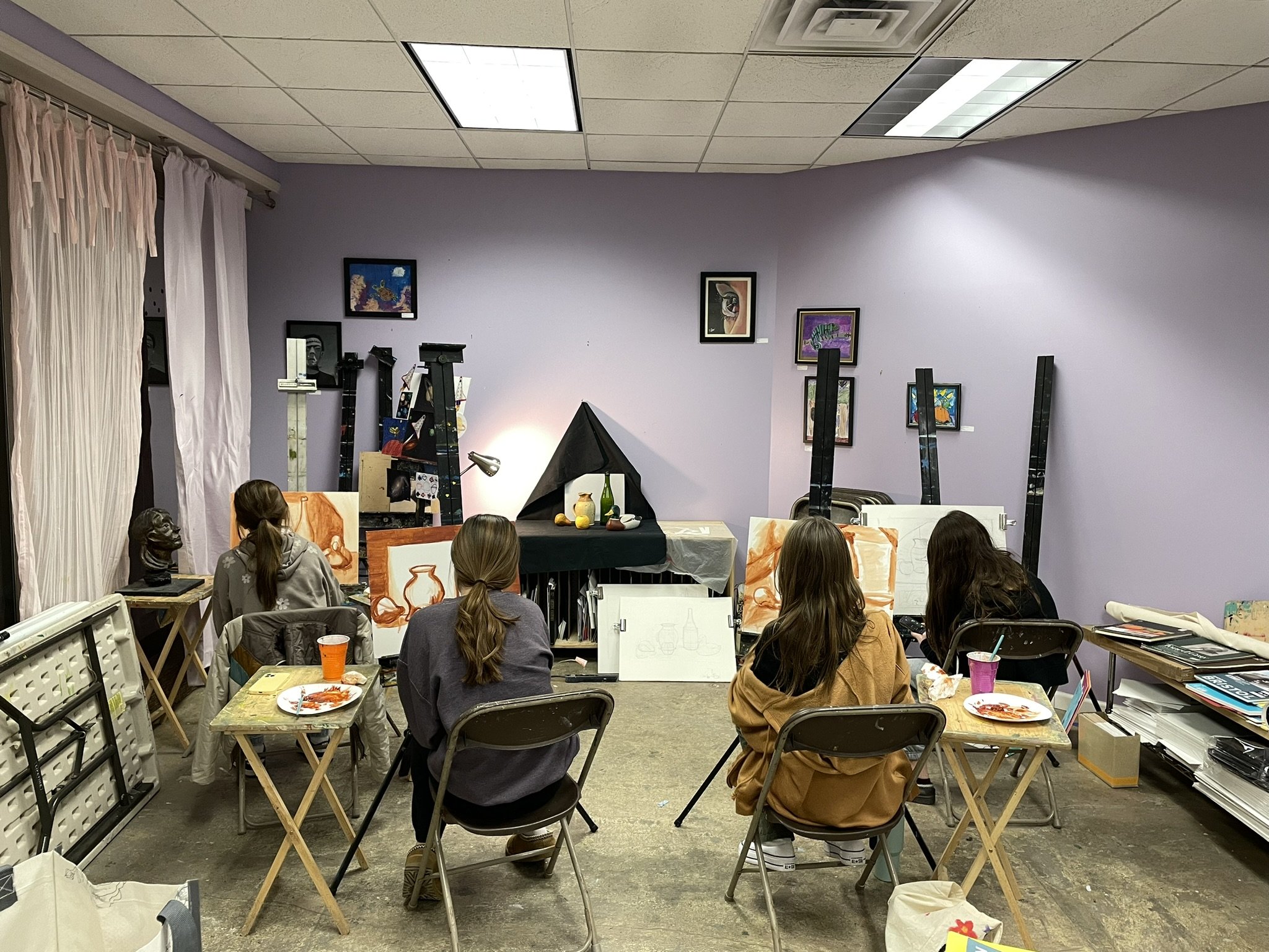 Four women sit at small tables in an art classroom working on paintings of vases. The room has purple walls decorated with framed artwork, art supplies, and easels.