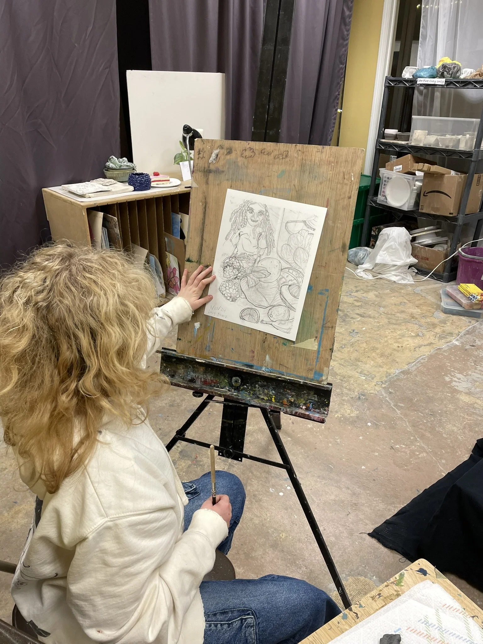 Child with curly blonde hair sitting at an easel sketching a still life with fruits. The studio has shelves and boxes with art supplies and storage.