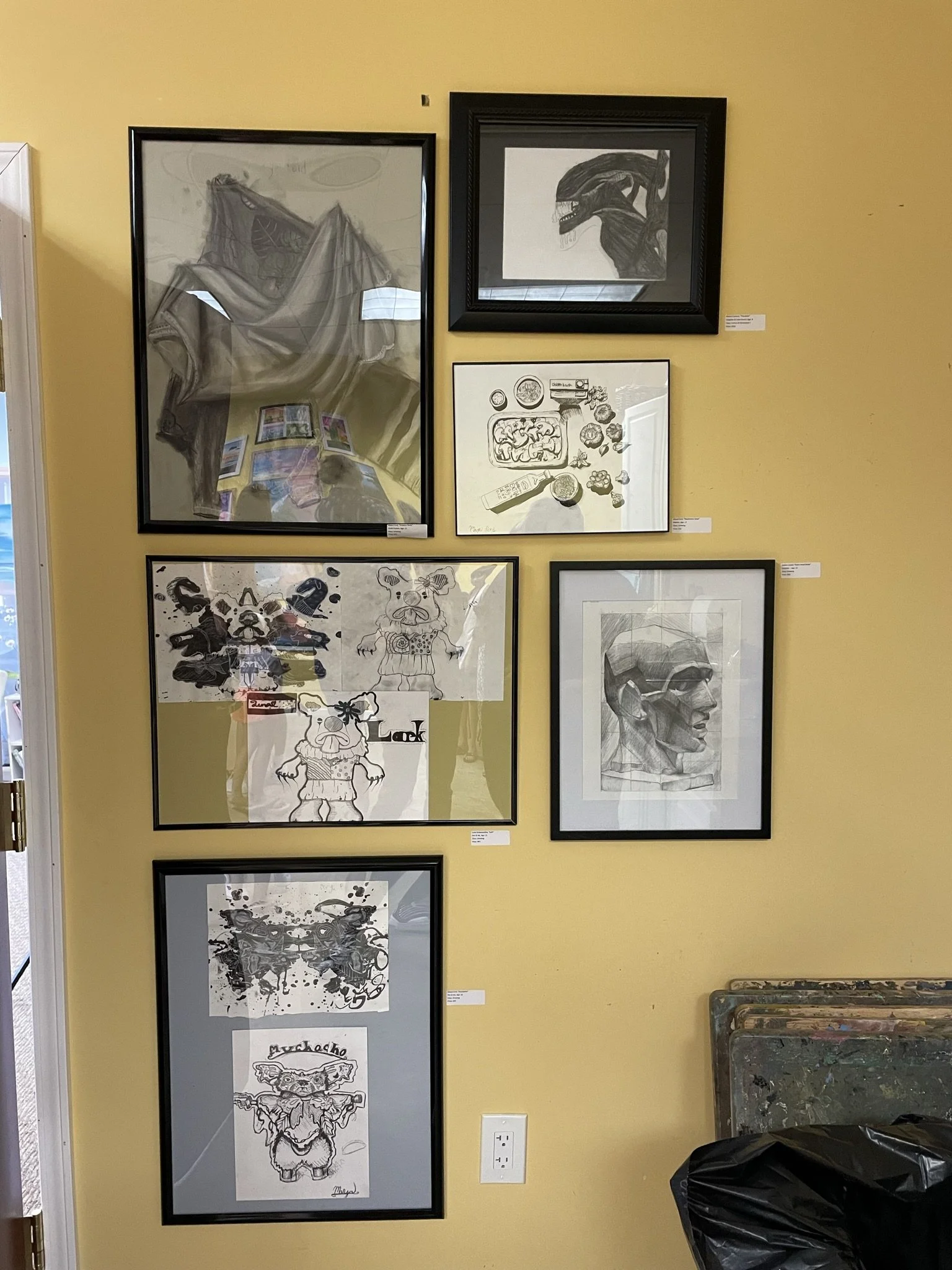 A yellow wall displays seven framed artworks, including black and white sketches, illustrations, and designs. Some frames contain abstract or cartoonish characters, while others feature detailed drawings of animal or creature heads.