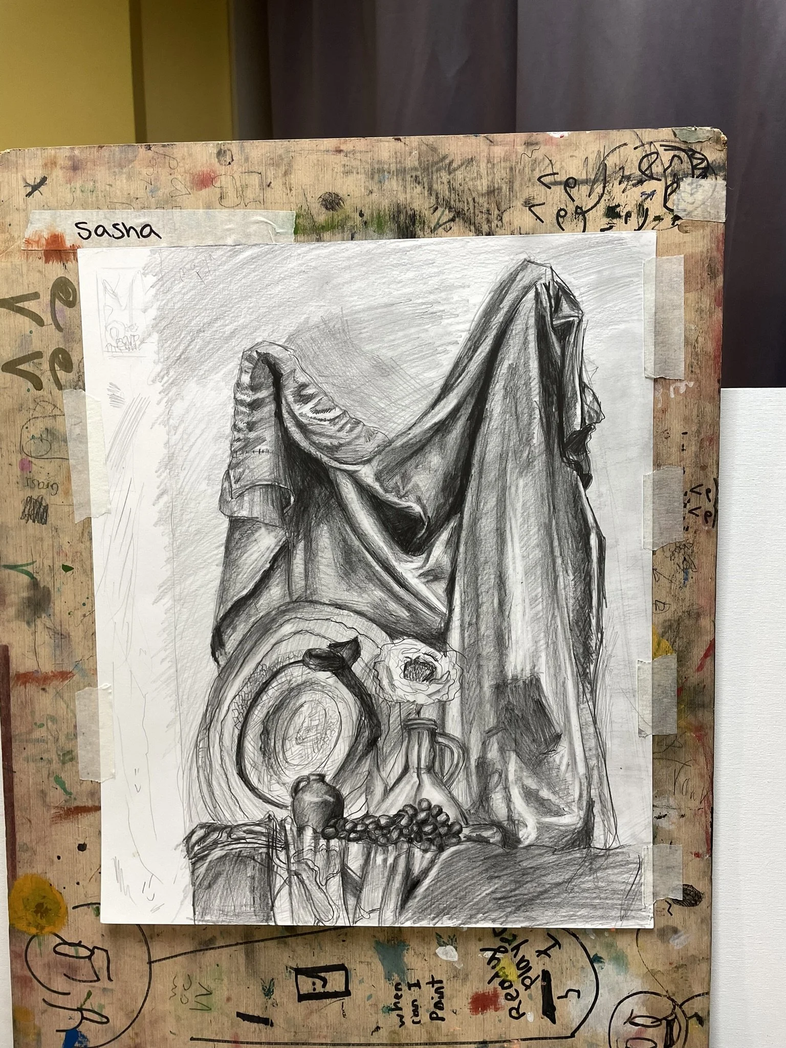 Black and white pencil drawing of a draped cloth, a hat, a flower in a vase, a bottle, and a string of beads. The drawing is taped to a paint-stained surface.