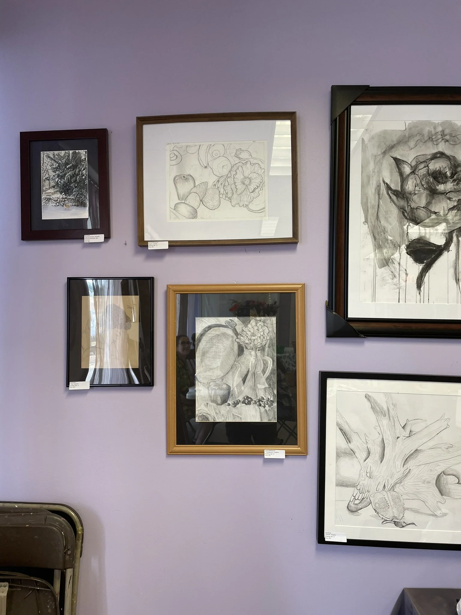 Collection of various framed artwork on a lavender wall, including sketches of a floral arrangement, pumpkins, and a tree stump with an acorn.