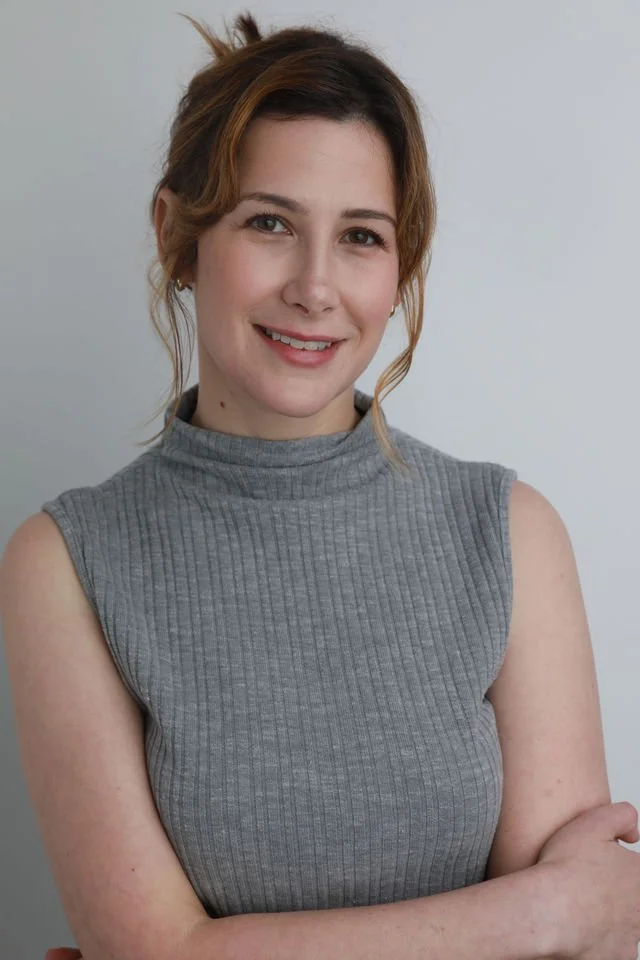 A young woman with light brown hair and a subtle smile, wearing a sleeveless gray ribbed turtleneck top, standing against a plain light gray background.