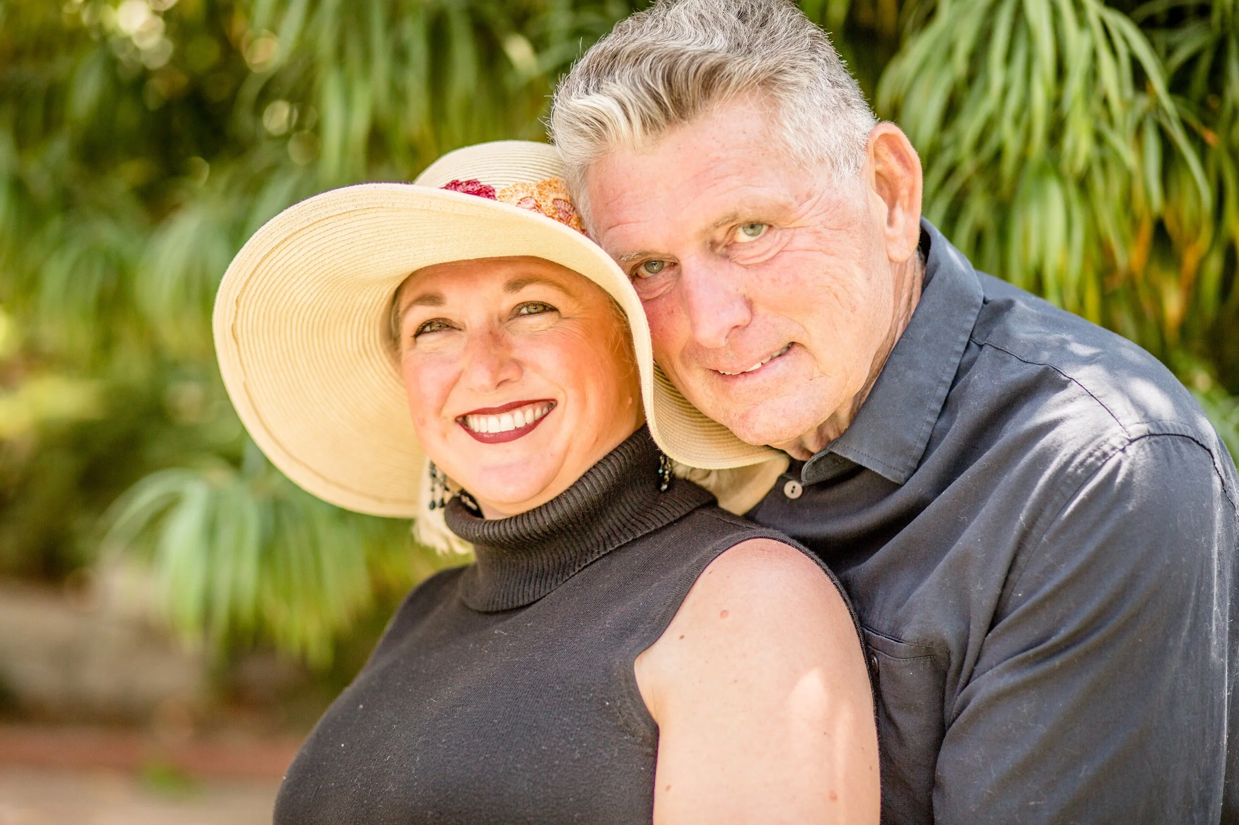 A smiling woman wearing a large sun hat and black sleeveless top, standing close to a man in a dark shirt outdoors with greenery in the background.