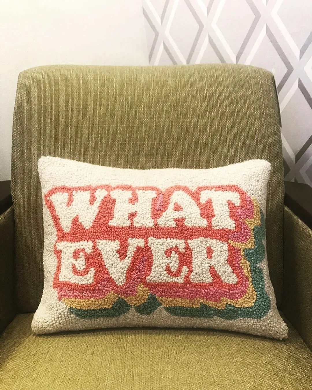 A square pillow with the words 'What Ever' in colorful, textured yarn, placed on a green upholstered chair.
