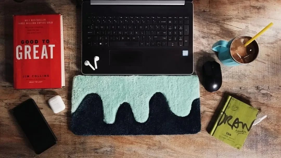 A flat lay of a wooden desk with a laptop, a pair of wireless earbuds, a smartphone, a white AirPods case, a black computer mouse, a Red book titled 'Good to Great' by Jim Collins, a green sketchbook with 'Dikan' on the cover, a cup of iced coffee with a yellow straw, and a pastel green and dark blue patterned cloth.